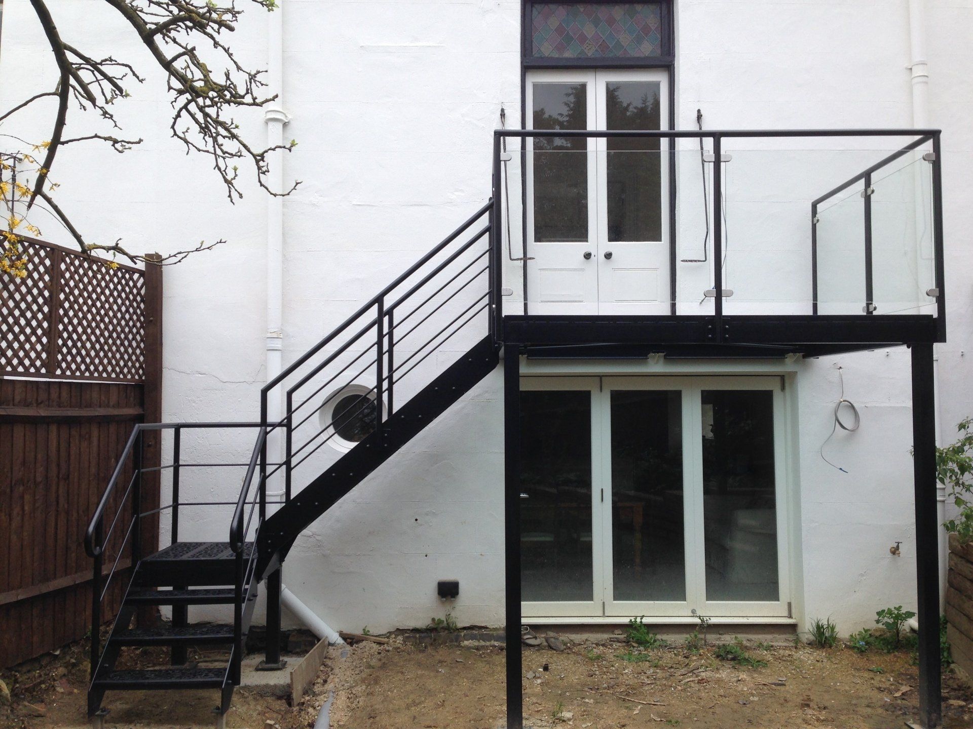 Black metal exterior staircase leading to a platform with glass railings. White building backdrop.