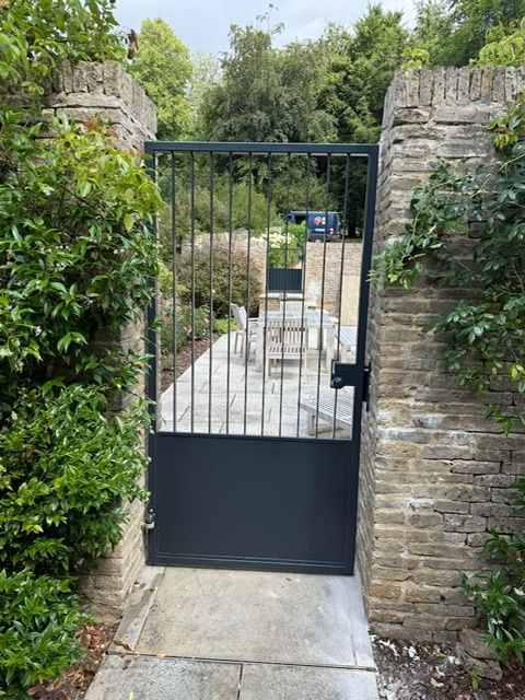 Black metal gate in a stone wall, leading to a garden path.