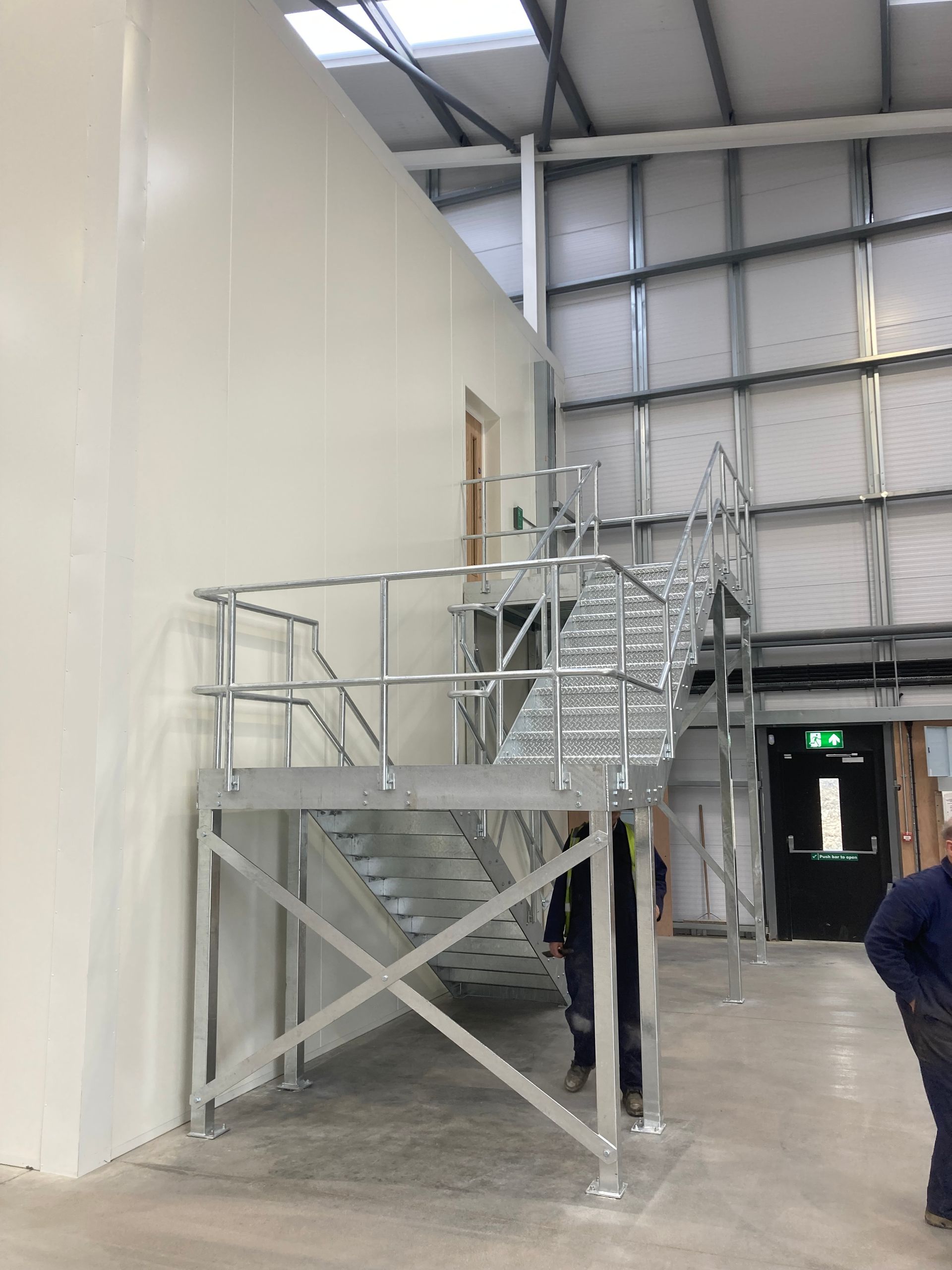 Metal staircase platform leading to a door inside a white building. Two people stand below.