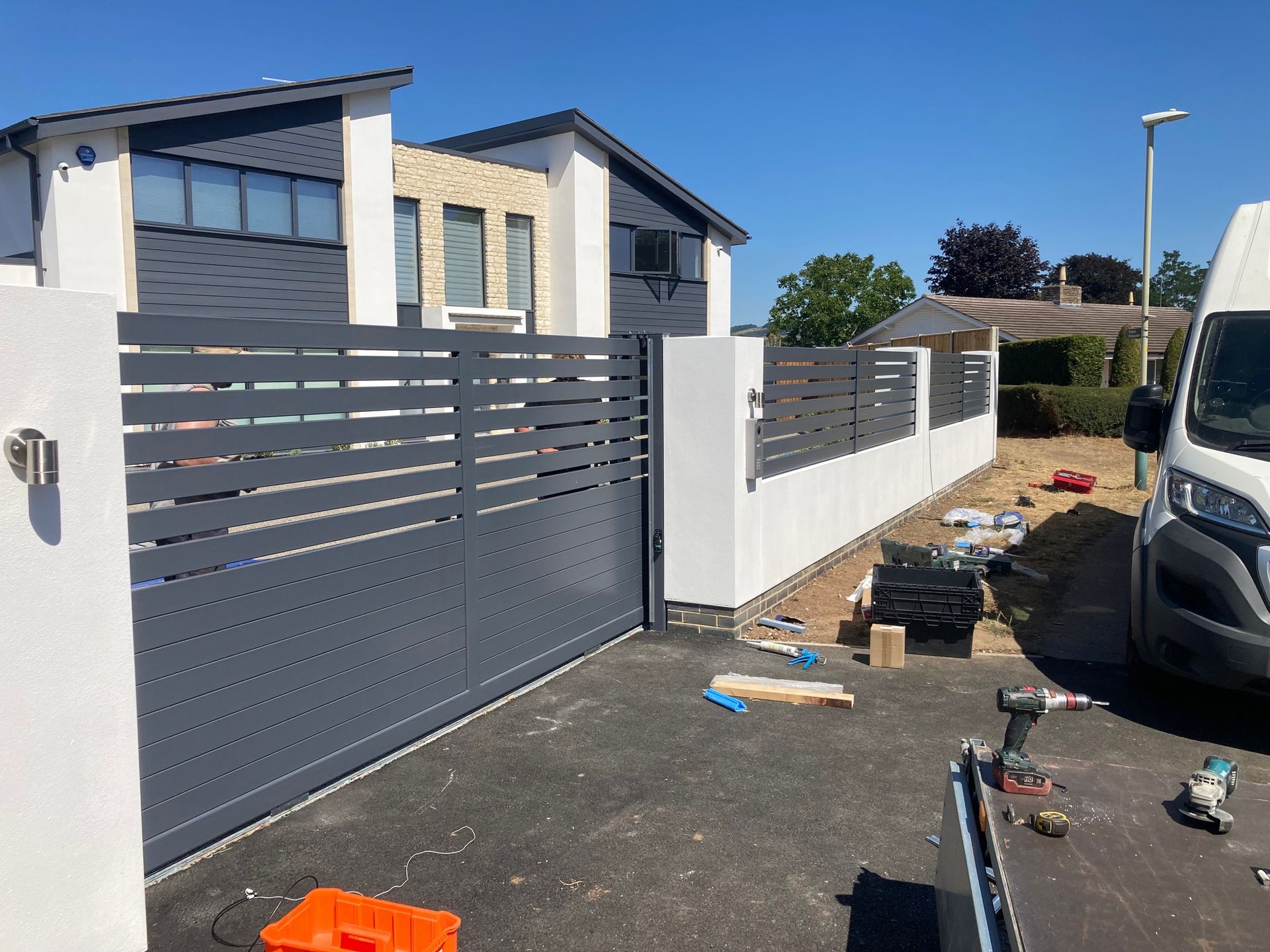 A gray horizontal slat gate and fence in front of a modern white house under a blue sky; construction tools present.