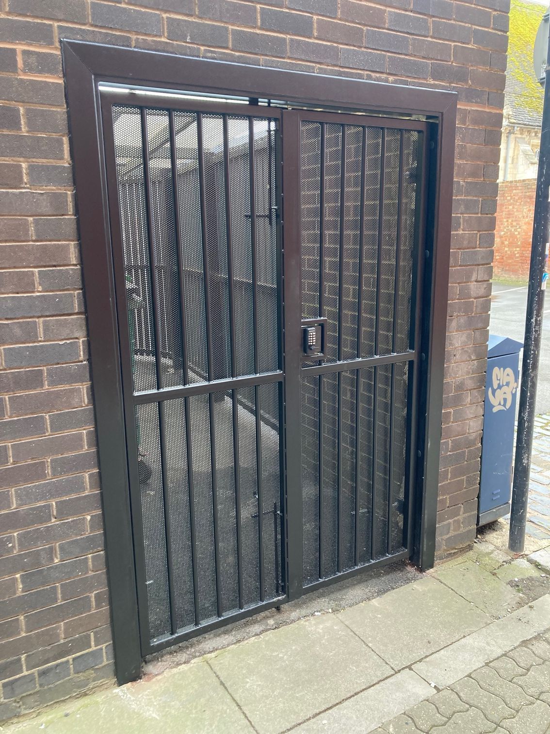 Black security gate in a brick doorway. Metal bars are vertical. Sidewalk and building exterior.