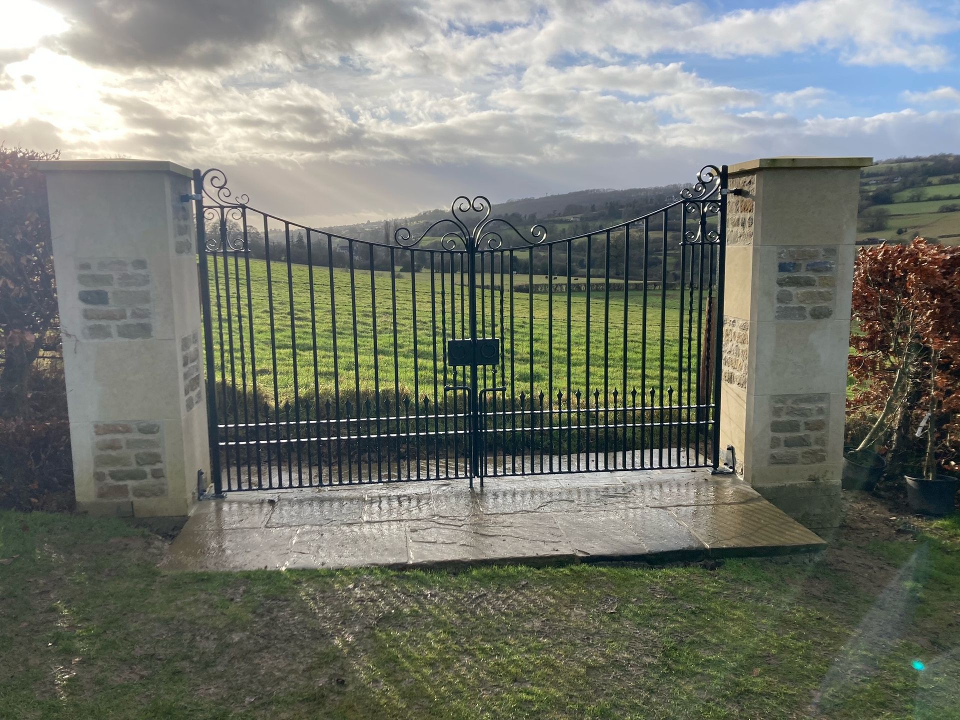 Black wrought-iron gate between stone pillars, overlooking a green field and hills under a cloudy sky.