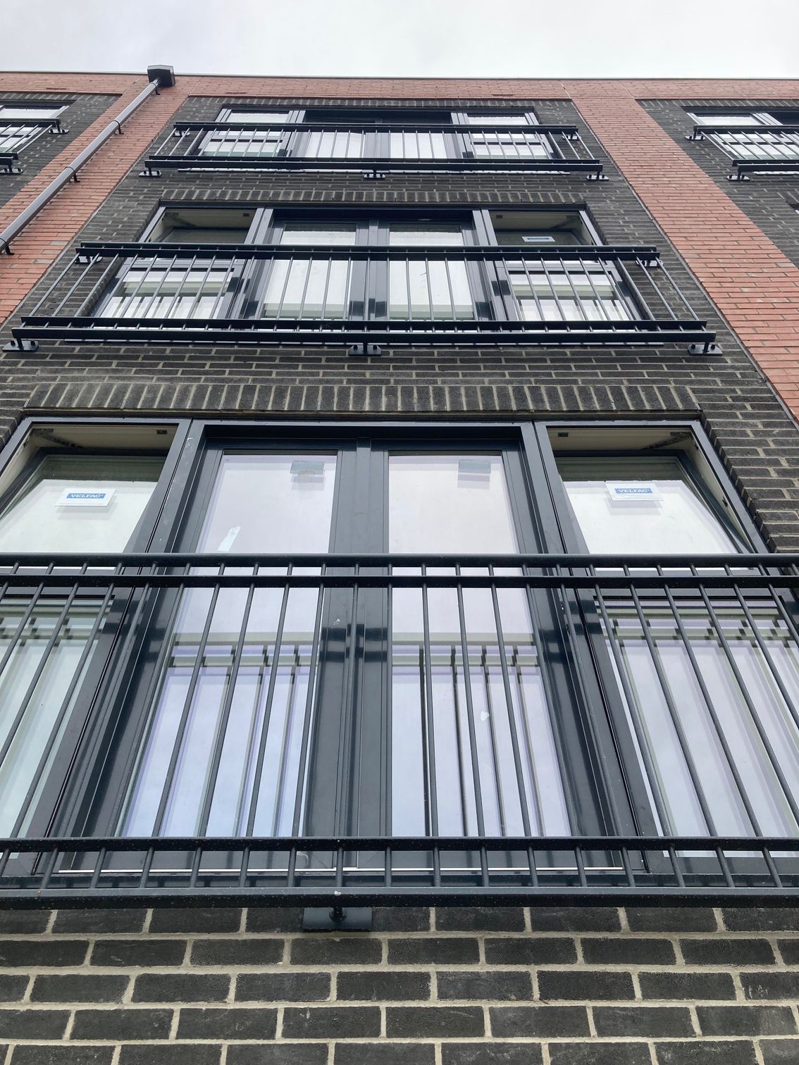 Low-angle view of a brick building with multiple balconies. Each balcony has a black metal railing and framed windows.