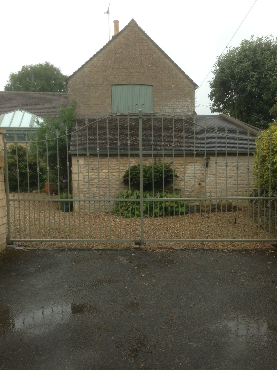 Stone building behind metal gate, gravel drive. Green double doors and small garden visible.