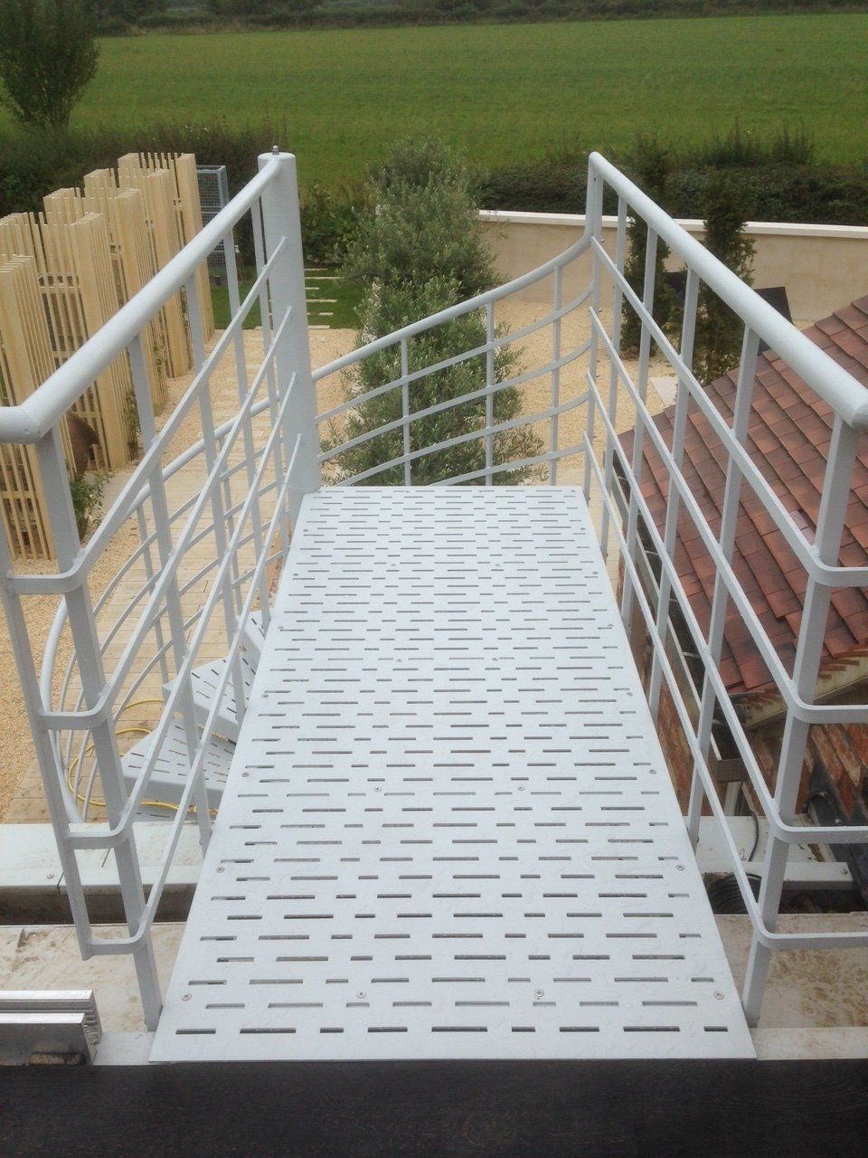 White metal ramp with railing and perforated walkway, outdoors with greenery.
