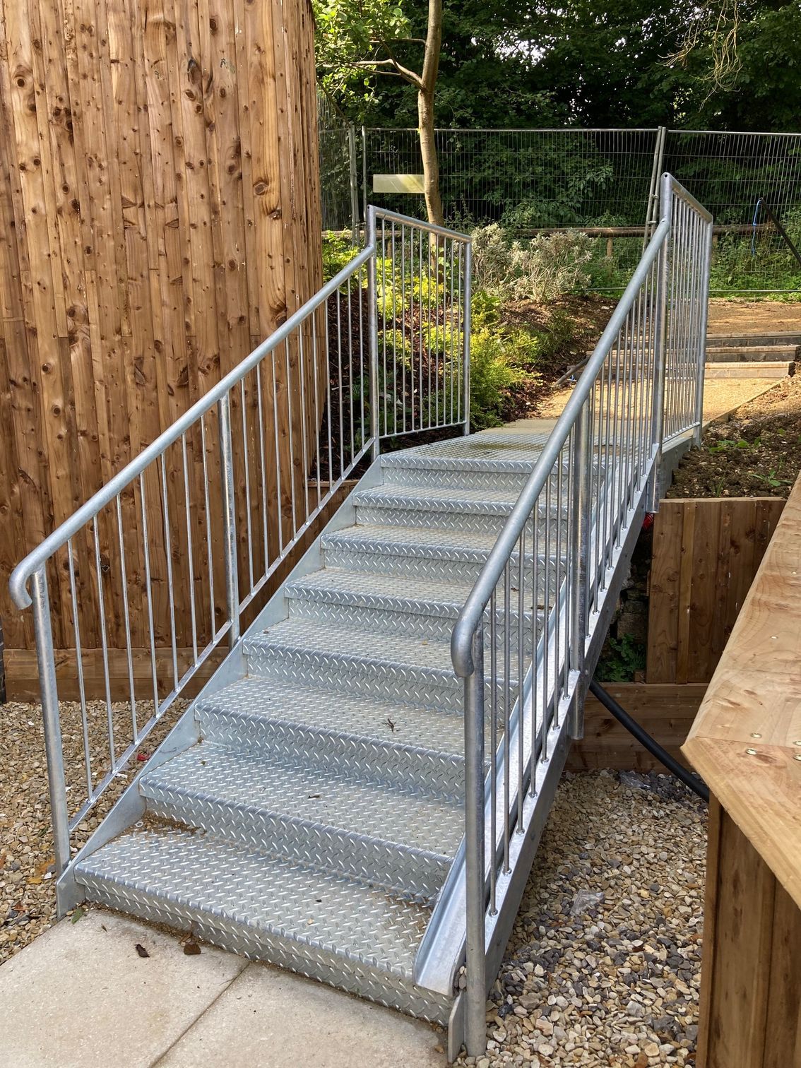Metal staircase with handrails, ascending from a concrete pad to a higher level with a wooden fence and foliage in the background.
