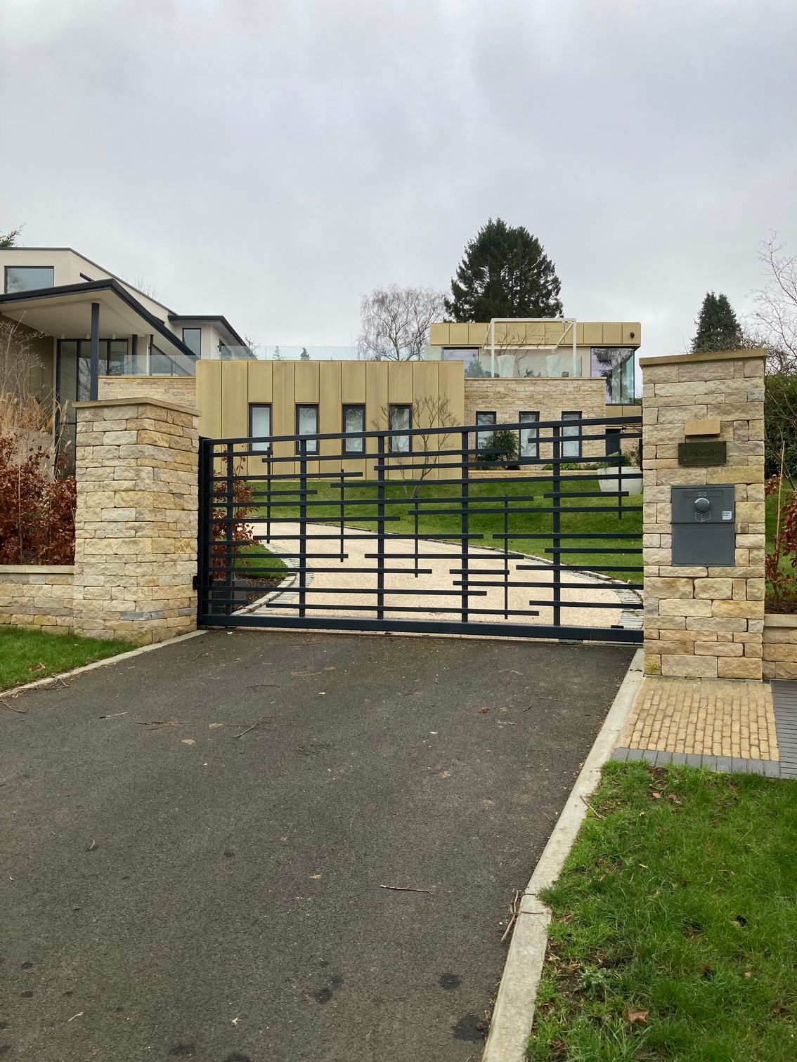 Black metal gate leading to a modern house with a beige facade. Stone pillars flank the gate on an asphalt driveway.
