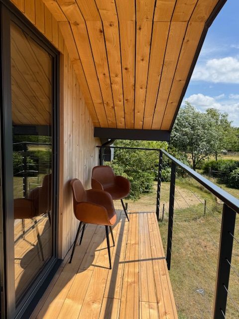 Balcony with wood siding, brown chairs, and black railing; sunny outdoor view.