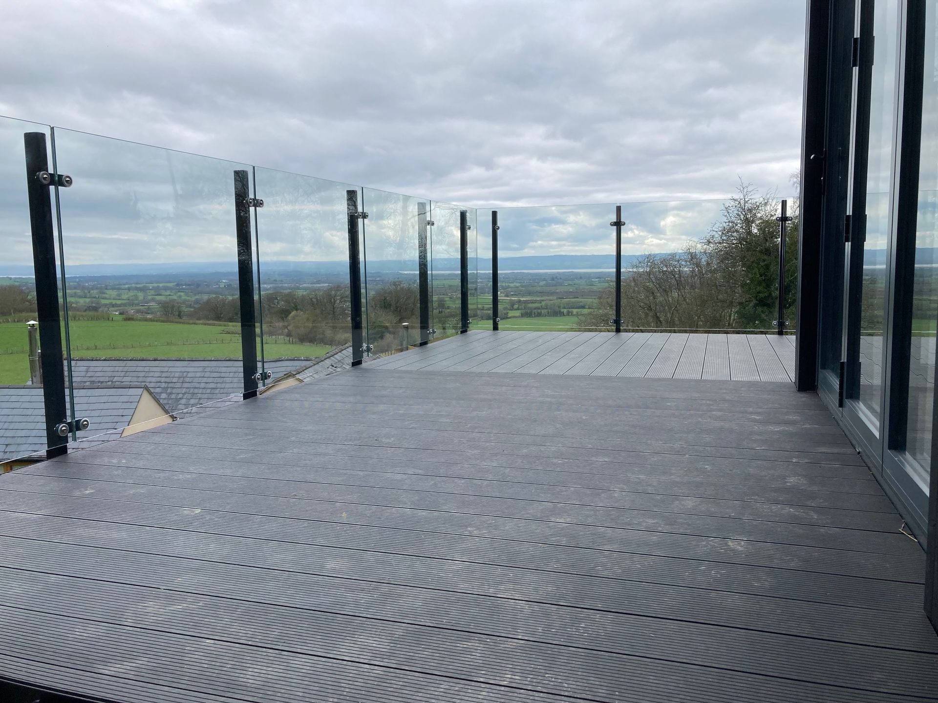 Grey composite deck with glass railing, overlooking a green landscape under a cloudy sky.