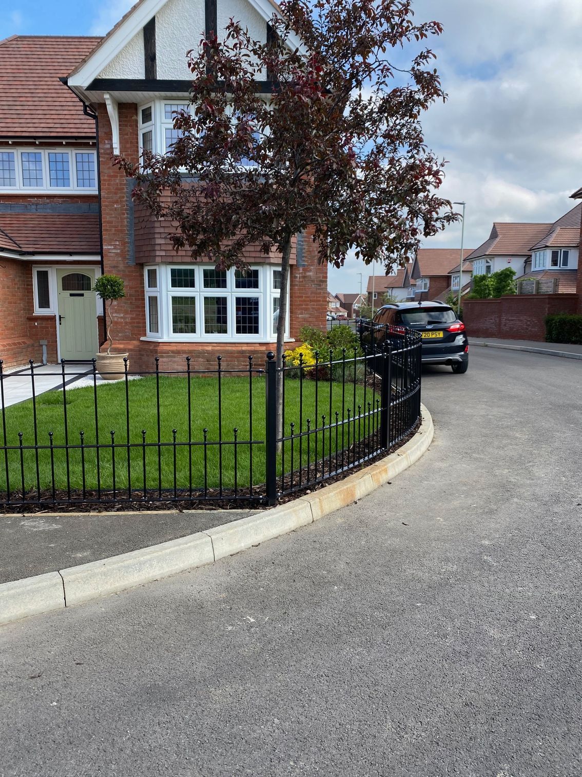 Black car parked on street, in front of a house with a small lawn, fenced with black iron.