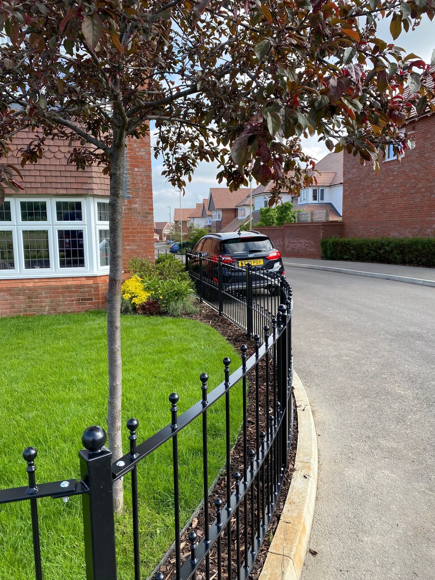 Black car parked on a curved residential street, behind a black wrought-iron fence.