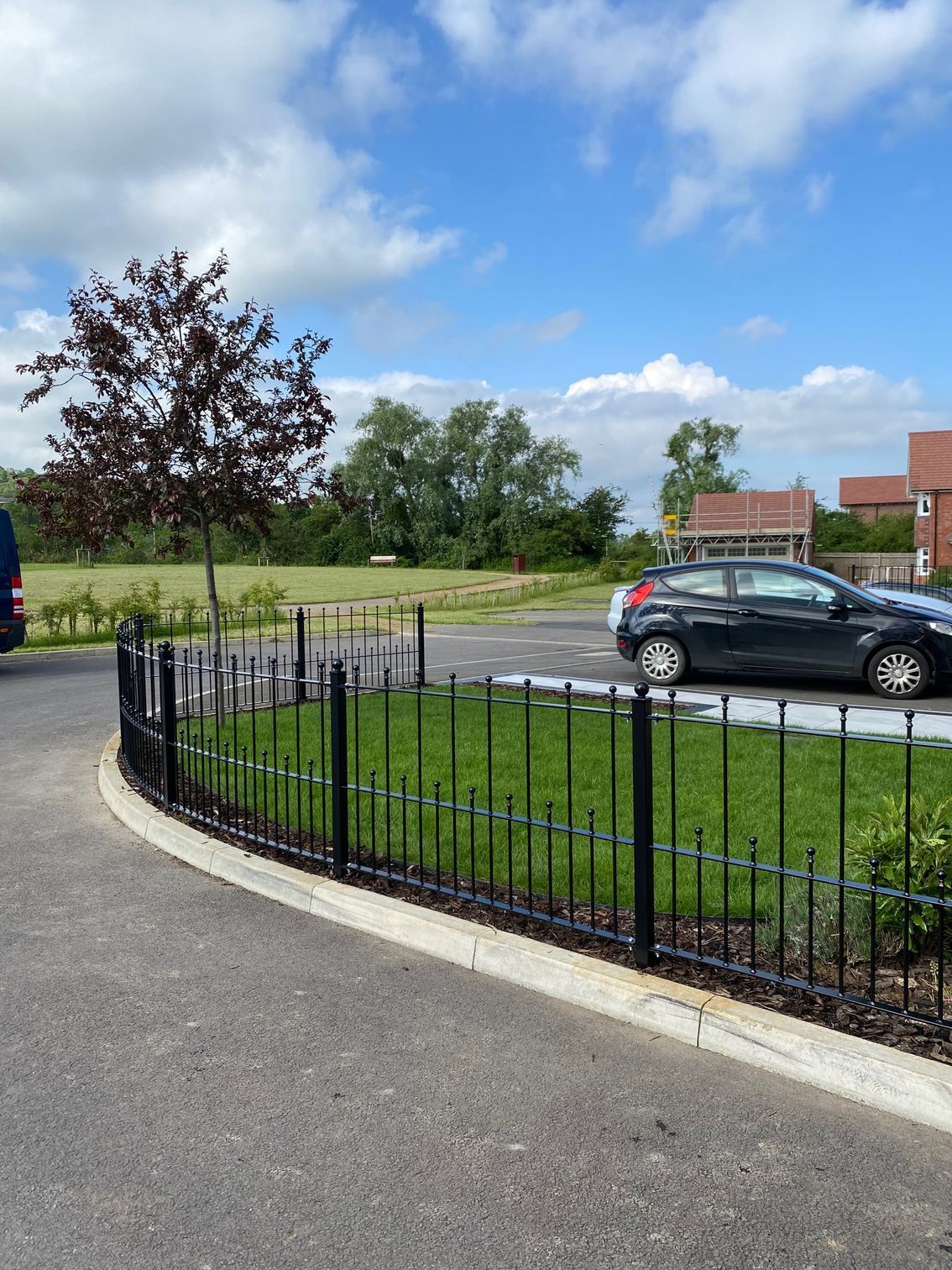 Black fence surrounds green grass, tree, and parked car on a sunny day.