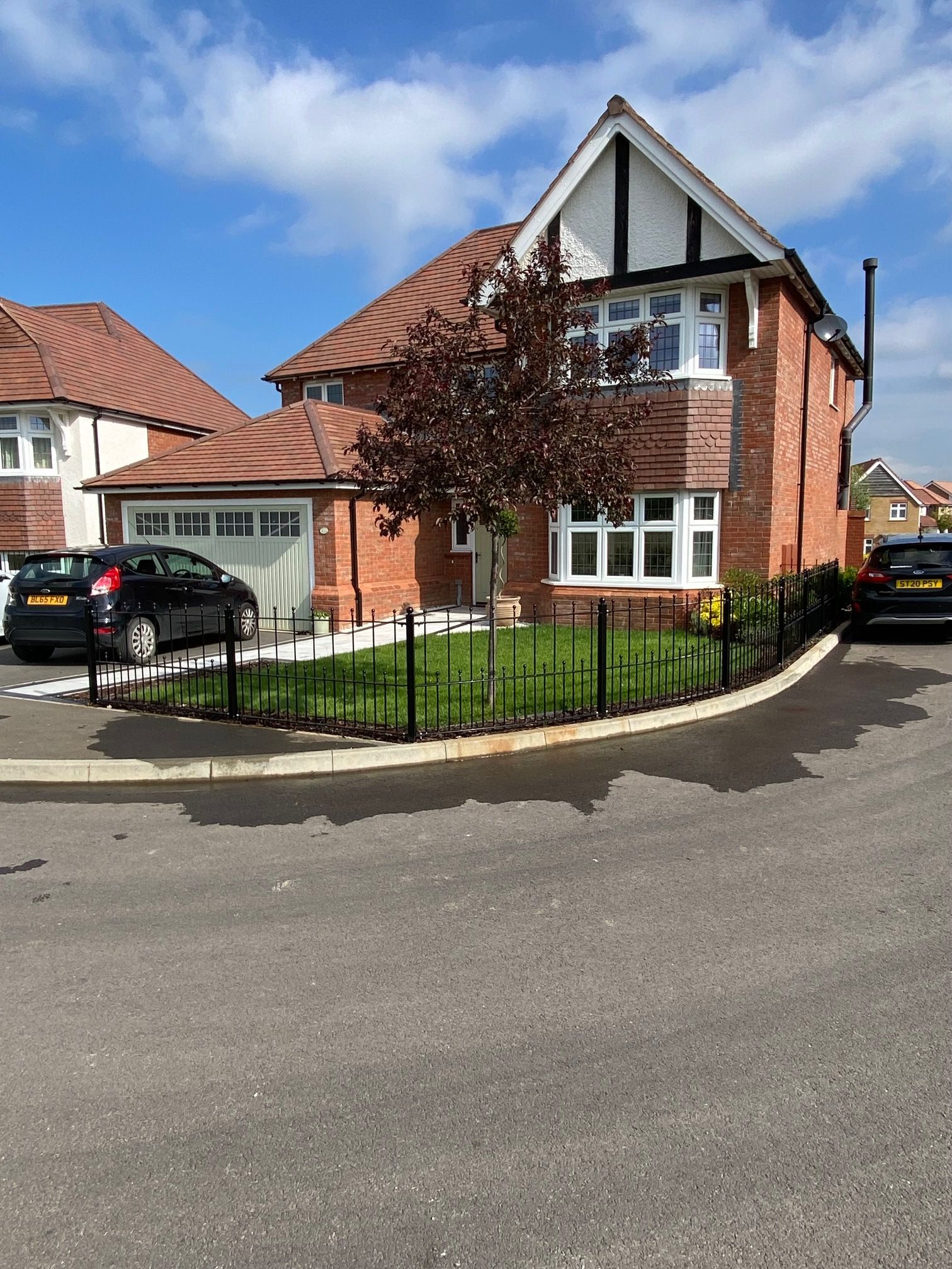 Brick house with a red roof, a small tree, black car, and a black fence.