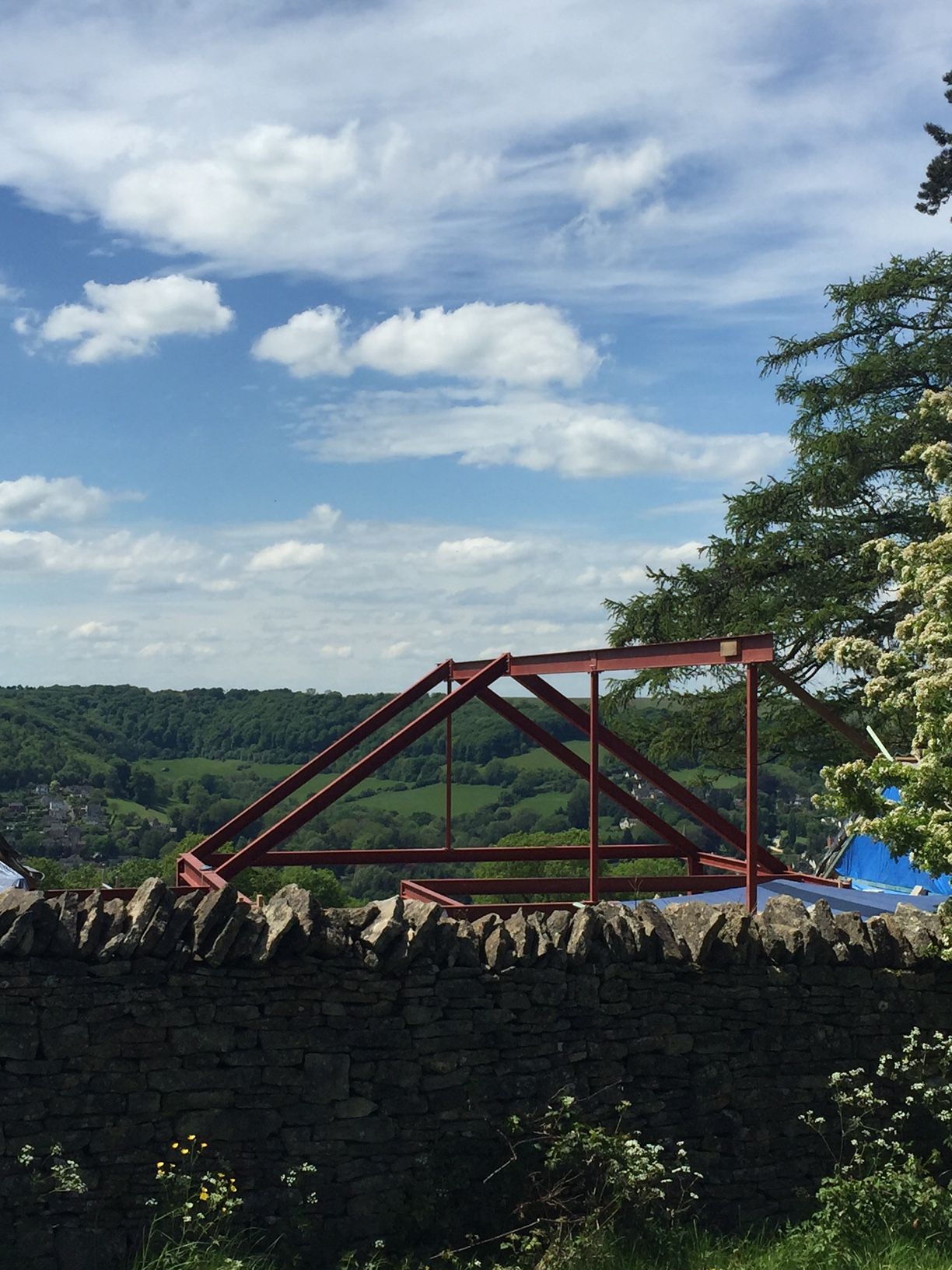 Red metal structure atop a stone wall, overlooking a green valley under a blue sky with fluffy clouds.