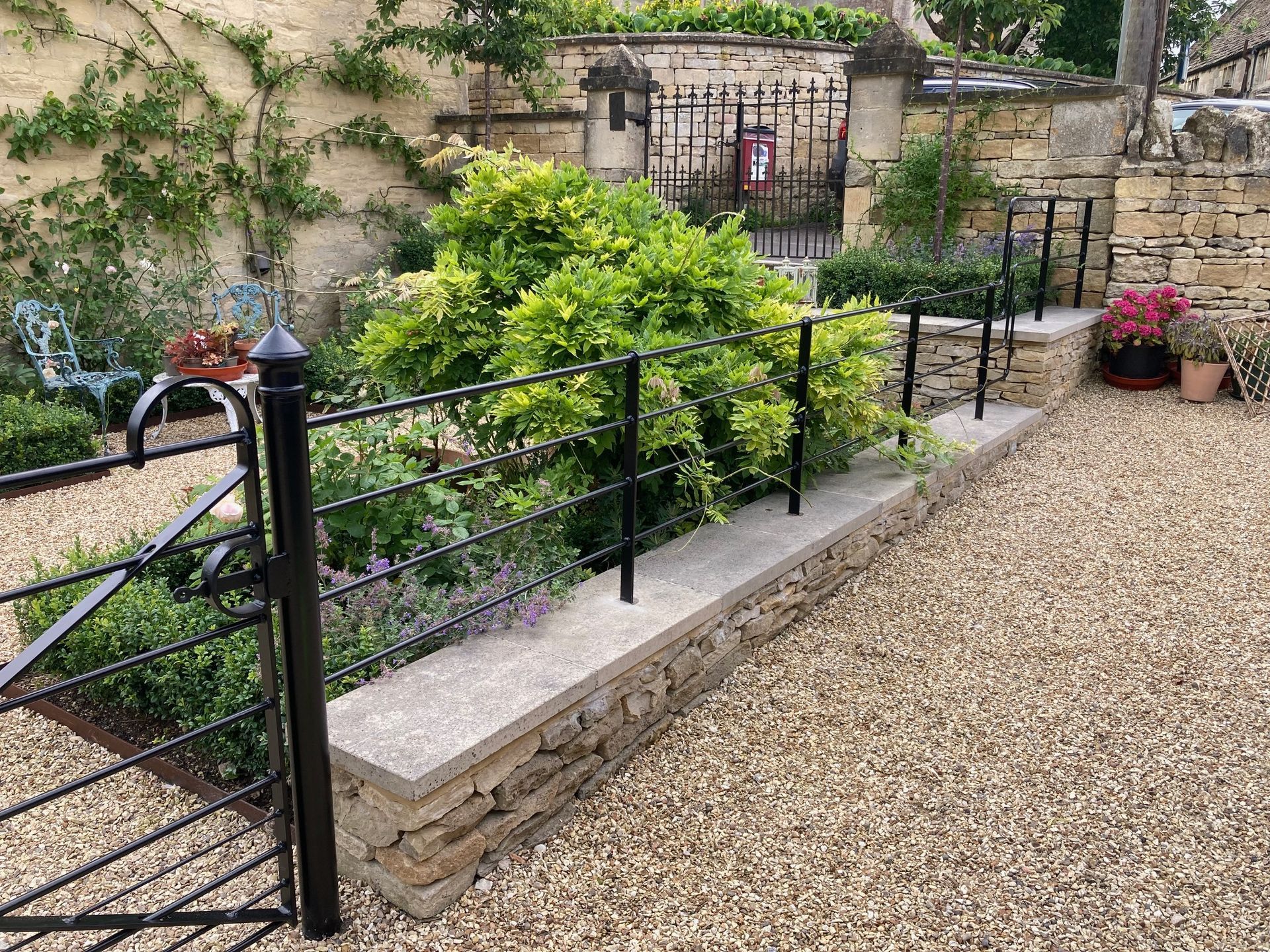 Stone wall with a black metal fence, gravel path, and greenery.