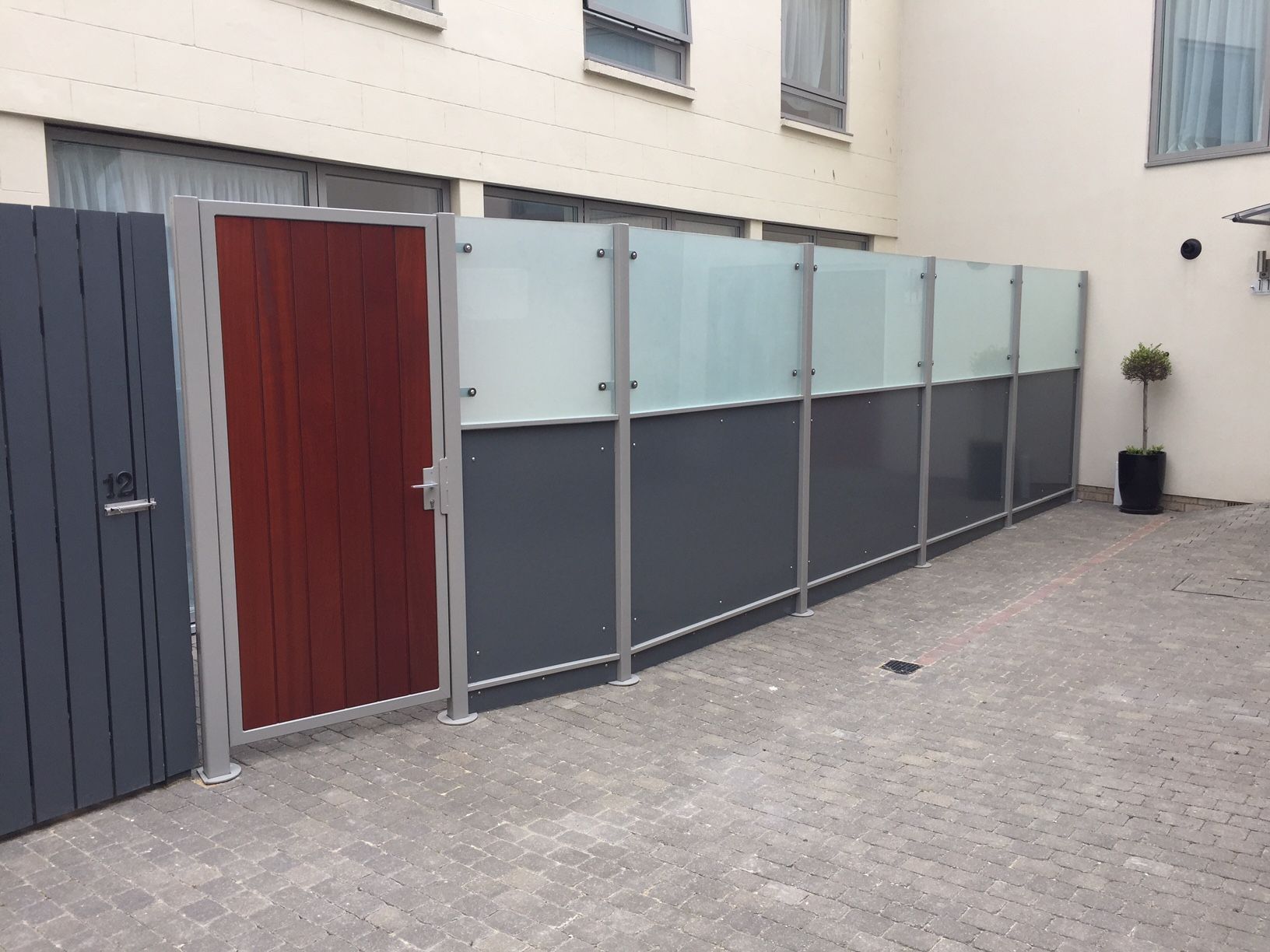 Gray and red fence with a frosted glass top section, enclosing a paved area.