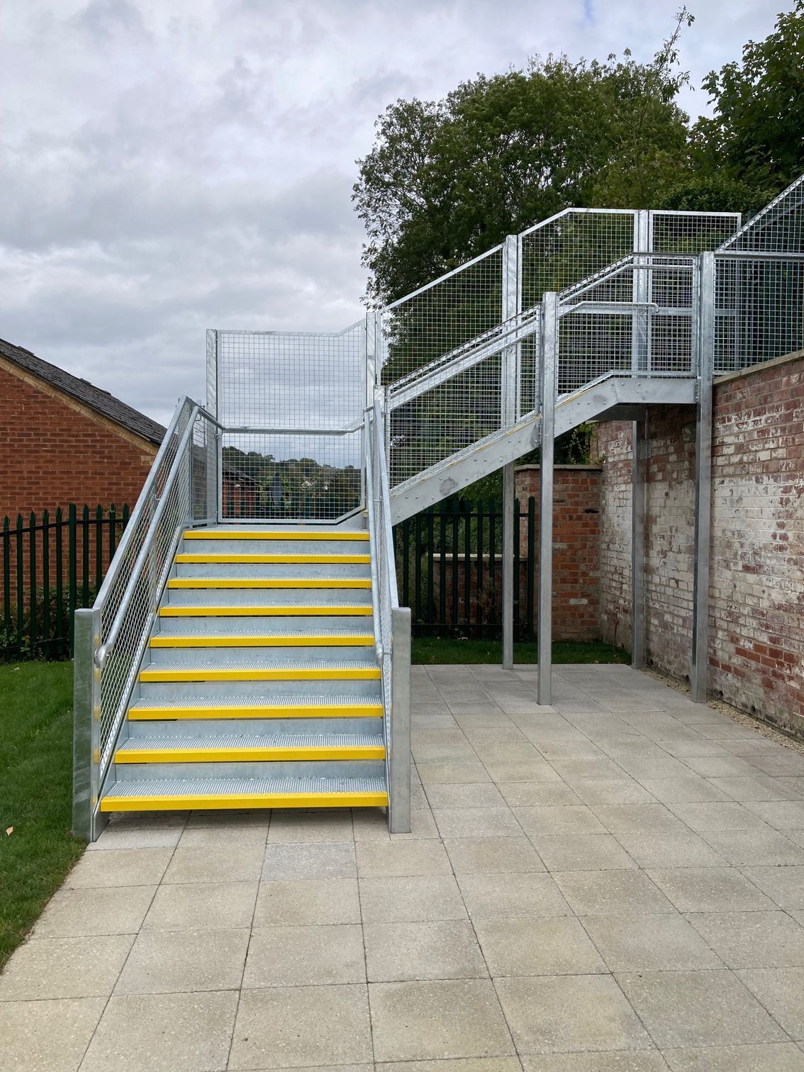 Metal outdoor staircase with yellow-edged steps, safety railings, and wire mesh fencing, next to brick wall and grass.