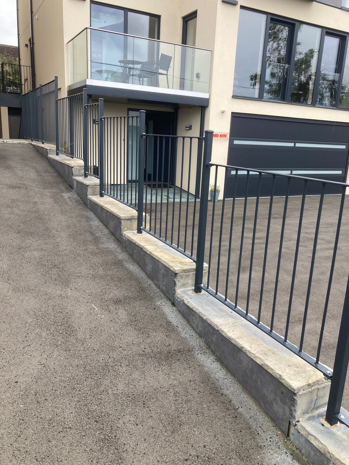 A driveway slopes up toward a modern house with a metal fence along a low concrete retaining wall.