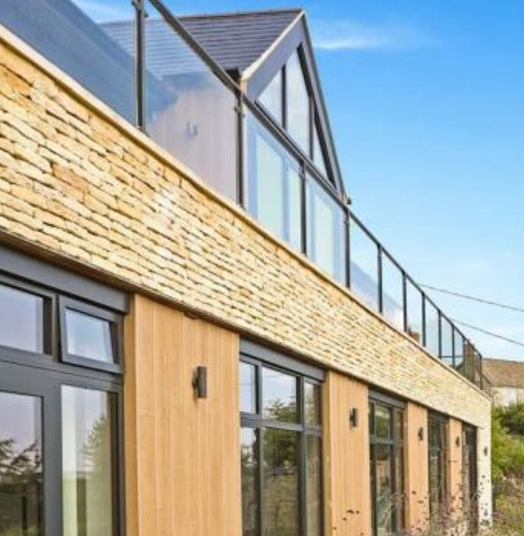 Modern Cotswold stone house with stone facade, glass balcony, and large windows against a blue sky.