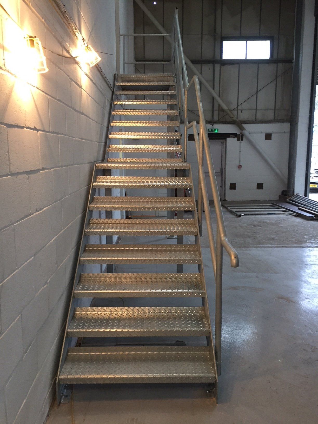 Metal staircase in an industrial setting, with handrail and wall lighting.