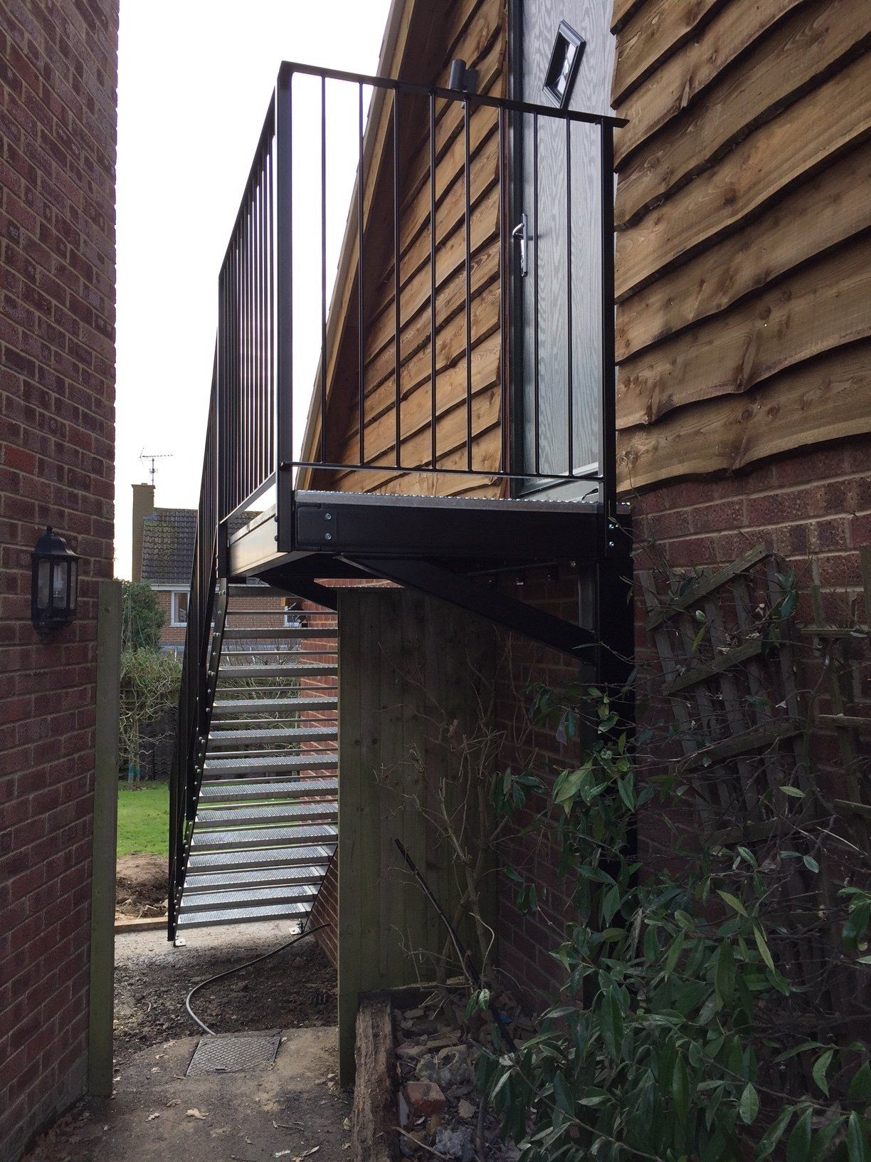 Metal staircase leading to a balcony on a wooden building, between brick walls and foliage.