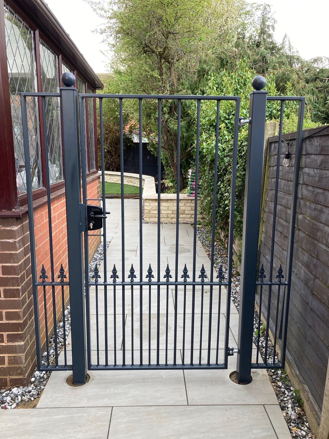 Black wrought iron garden gate on a stone path, leading to a yard, flanked by brick and a fence.