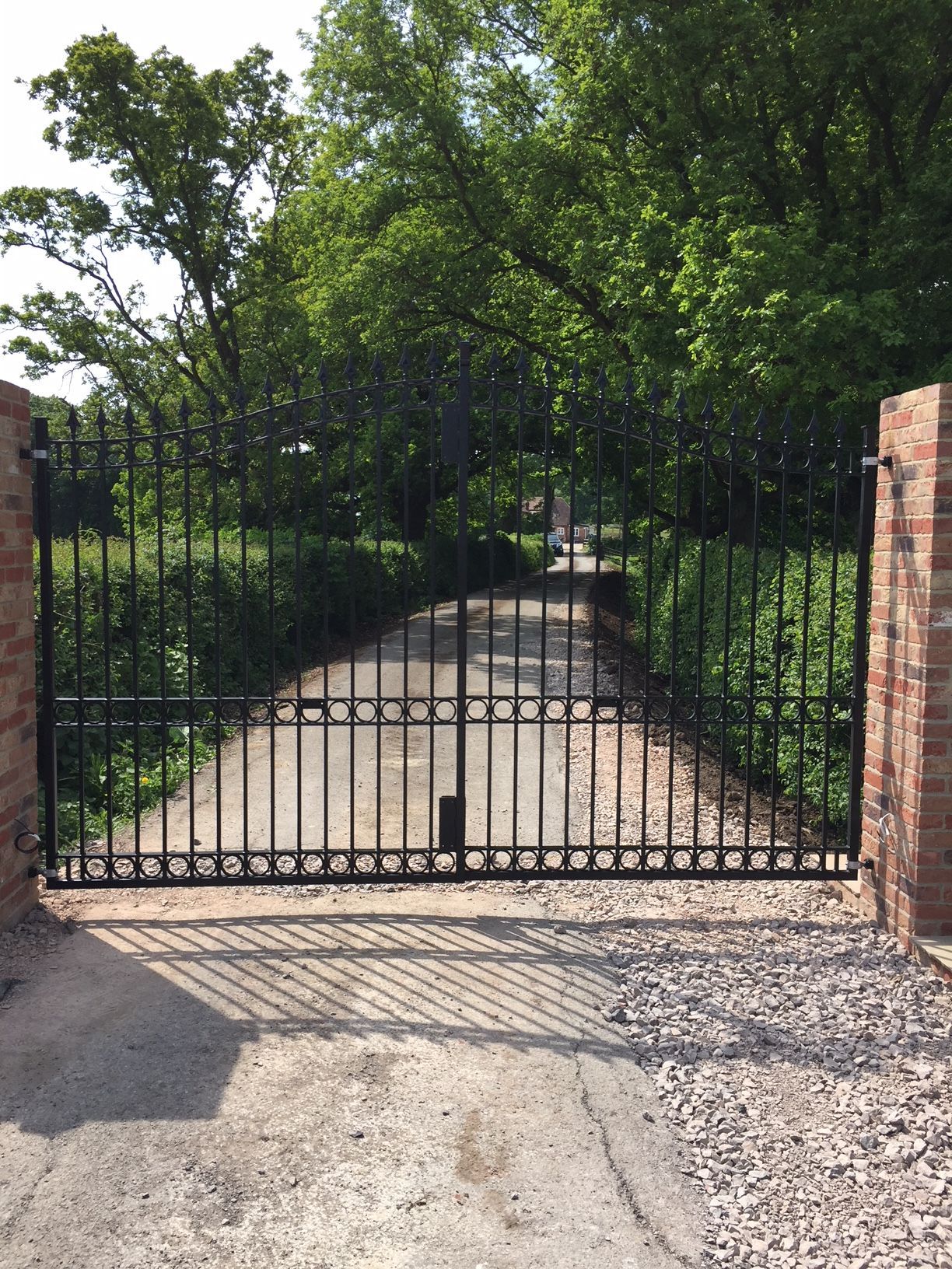 Black wrought iron gate set between brick pillars, guarding a gravel driveway, with trees in the background.