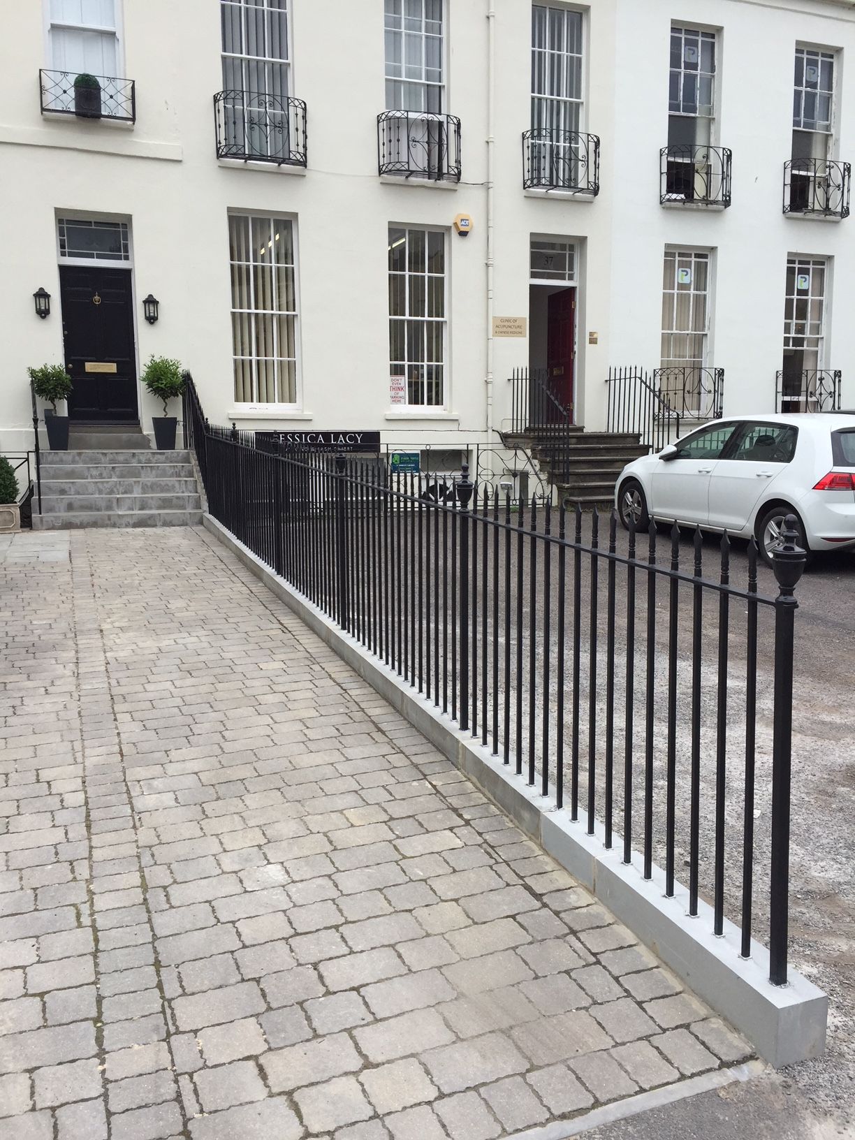 Black metal fence bordering a paved driveway leading to a white building with windows and steps.