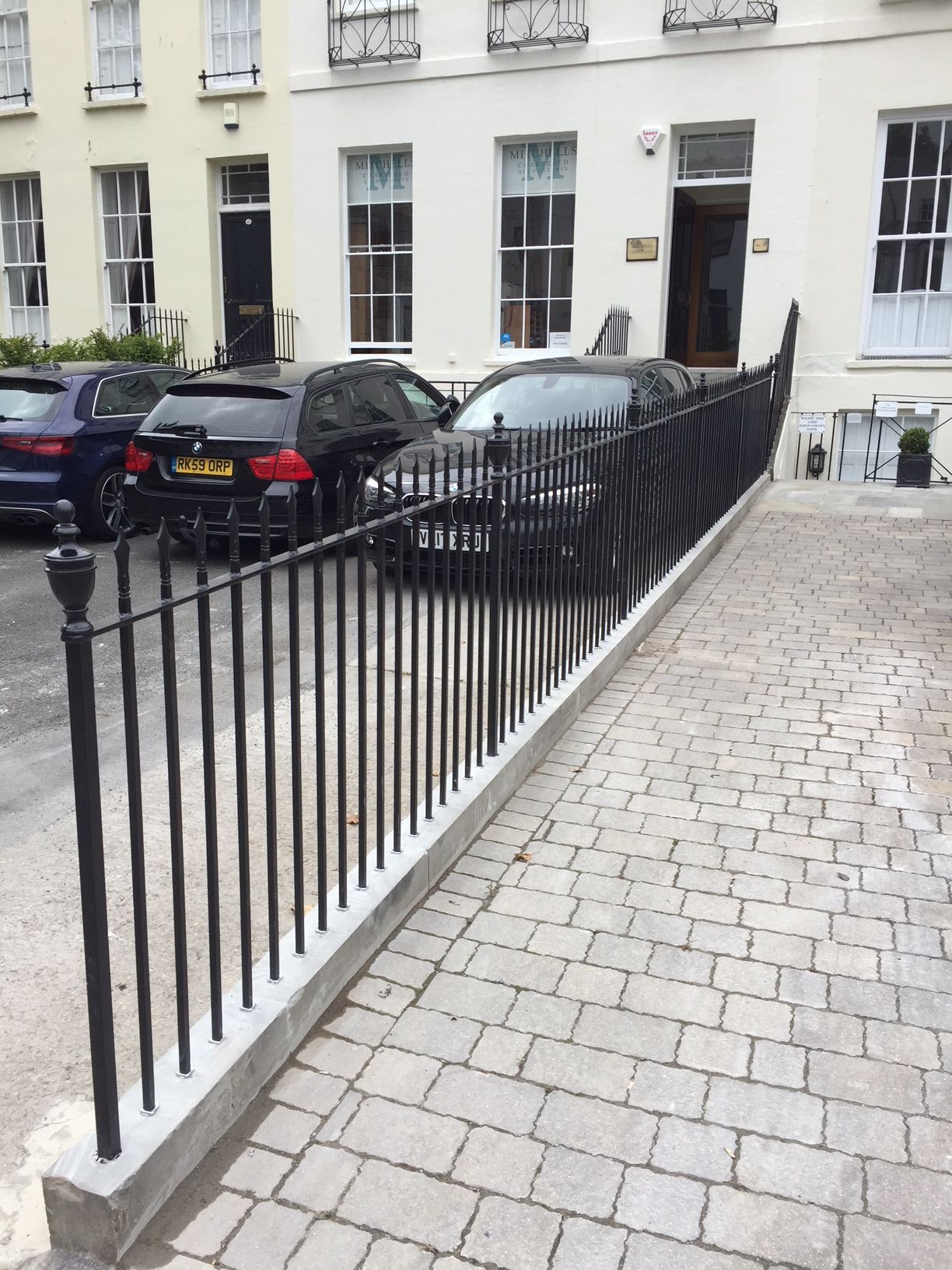 Black metal fence on a concrete base, separating a paved area from parked cars in front of a building.