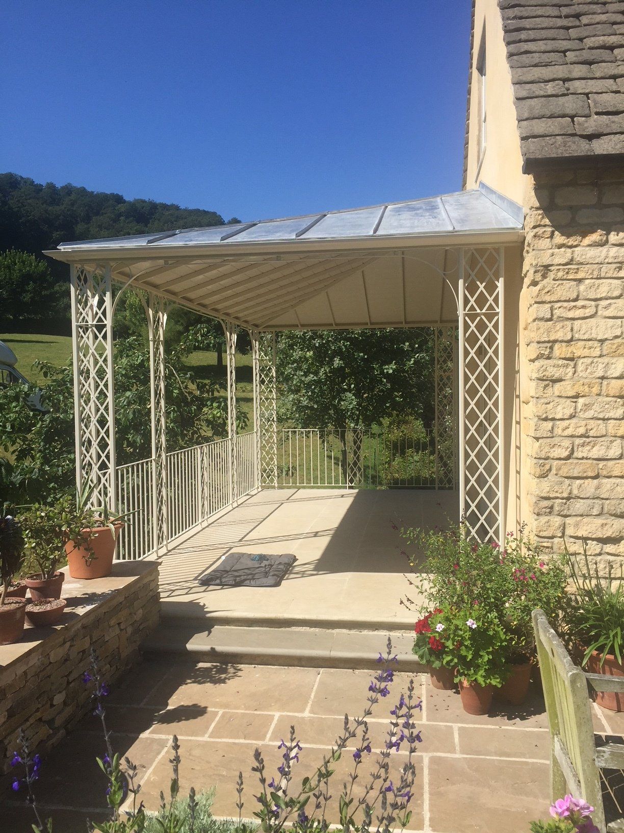Covered porch with white metalwork, stone steps, and potted plants. Sunny day.