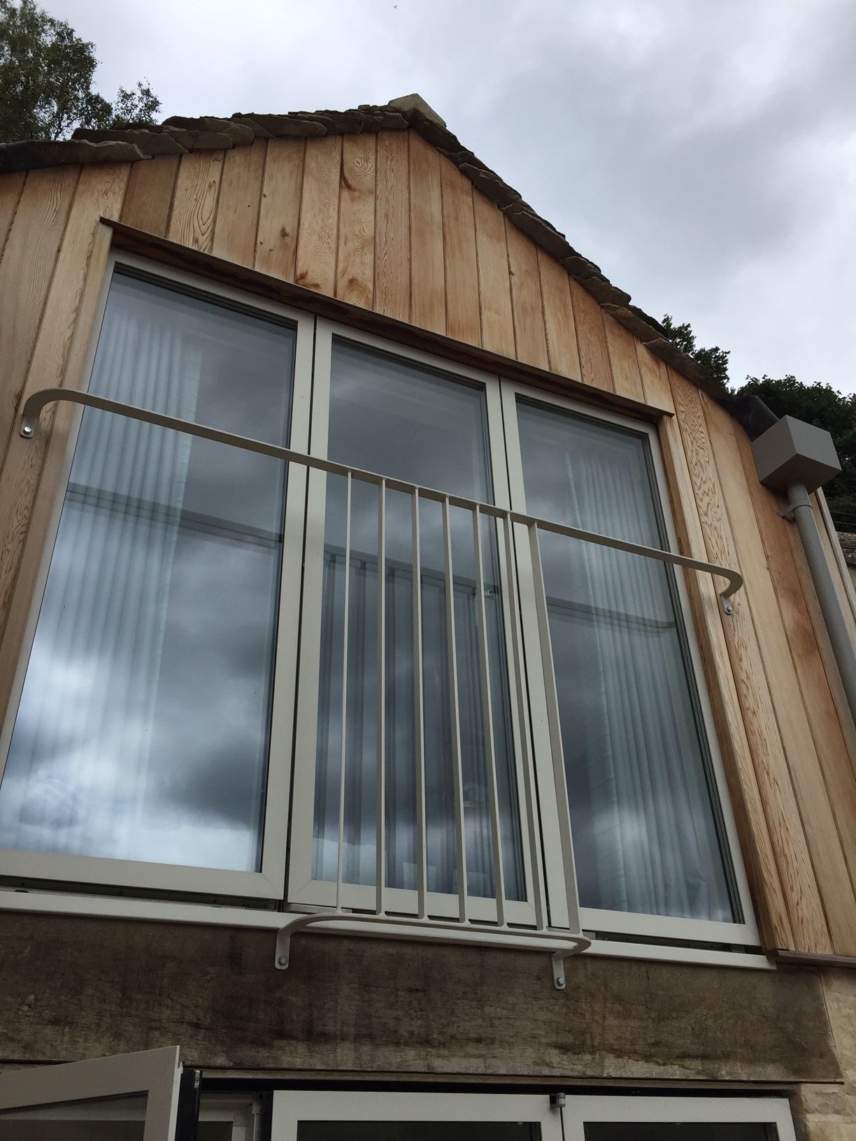 Exterior view of a building with large windows and a small balcony railing. Wooden siding. Overcast sky.