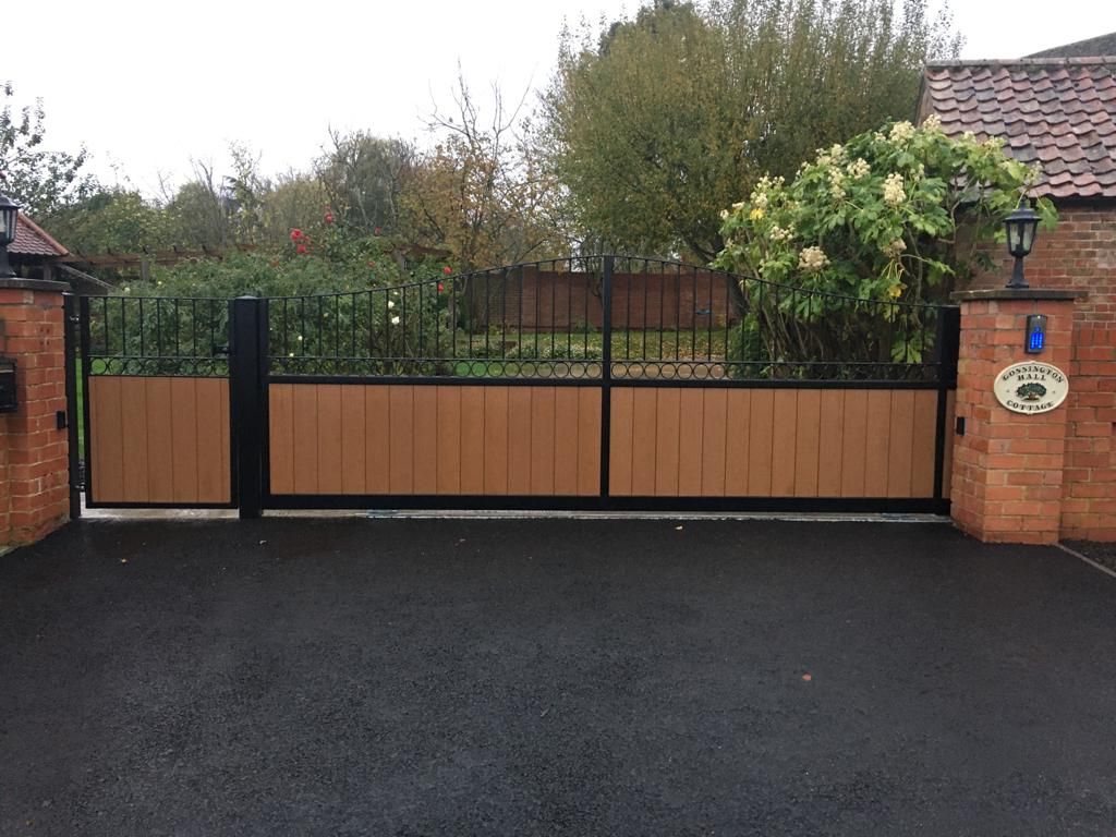 Brown and black driveway gate with wooden panels, brick pillars, and asphalt driveway.