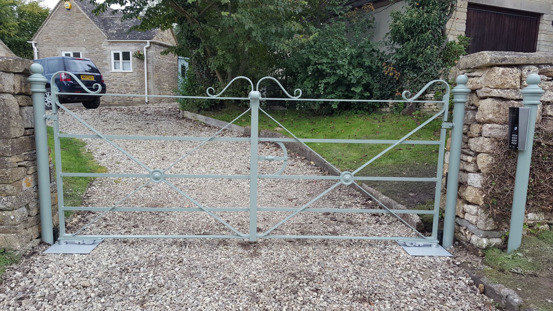 Gray metal gate across a gravel driveway, between stone pillars, leading to a house.