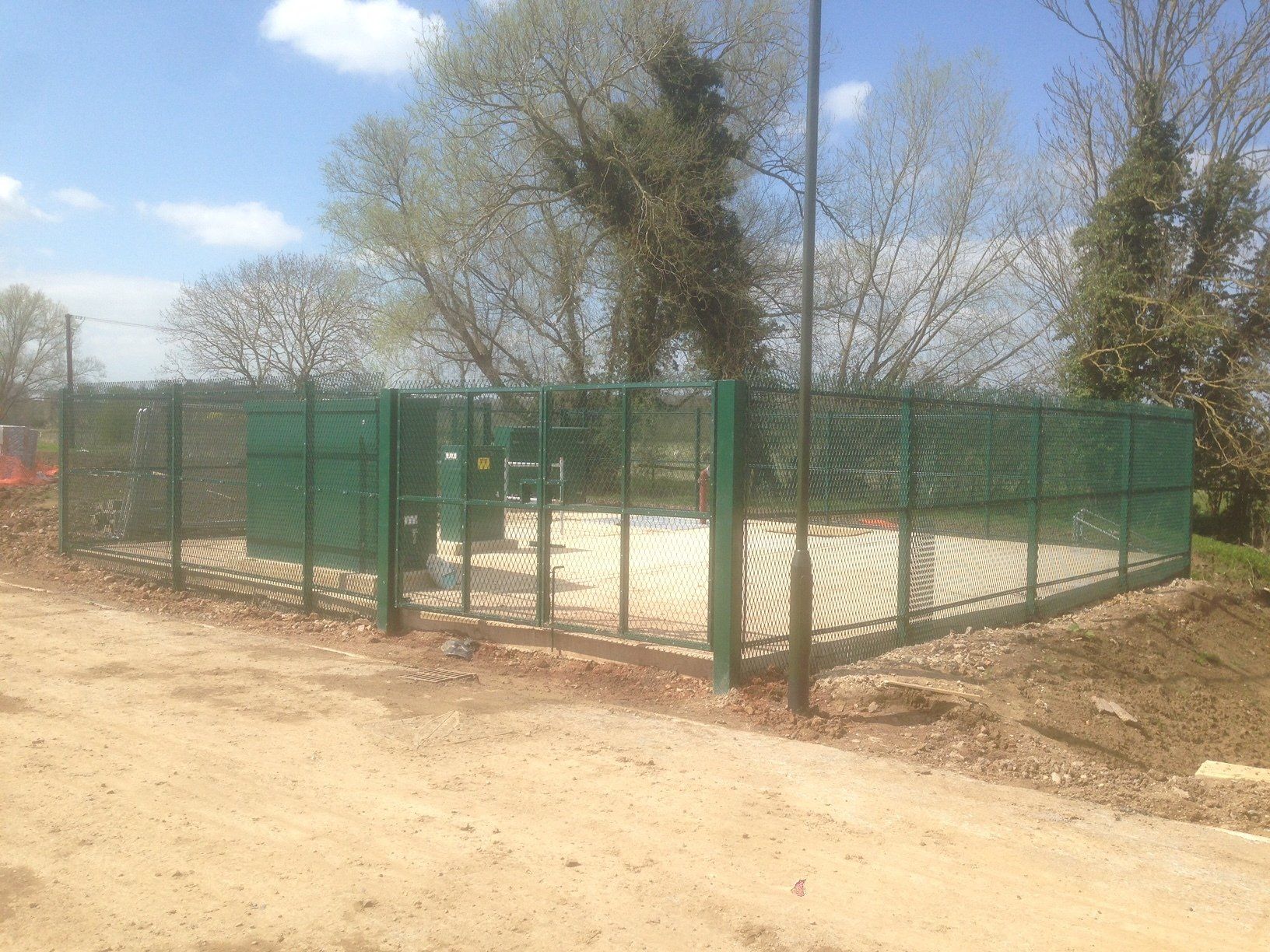 Green fenced utility area with equipment, set on dirt road under a sunny sky.