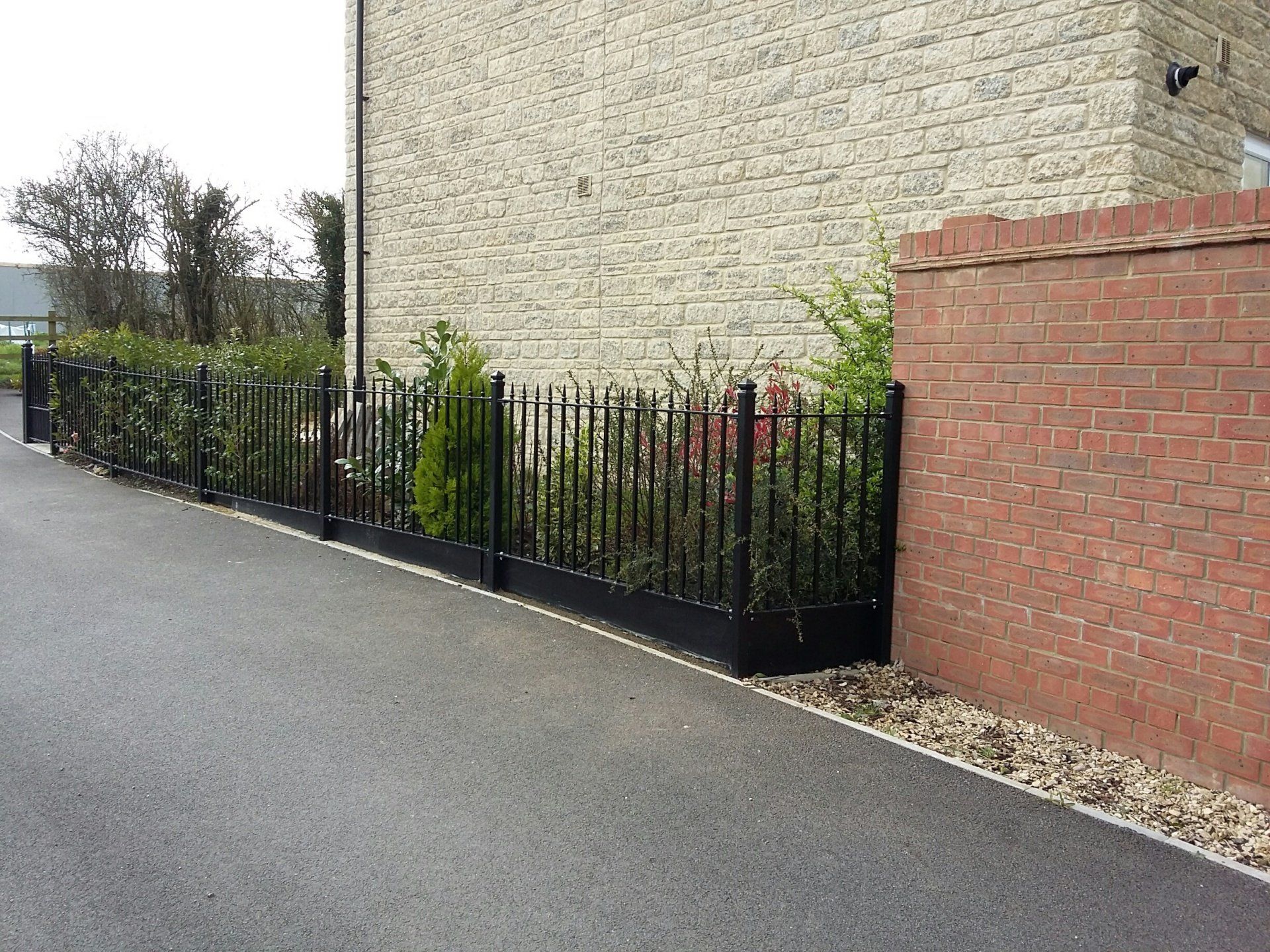 Black wrought iron fence with shrubs alongside a brick building and paved driveway.
