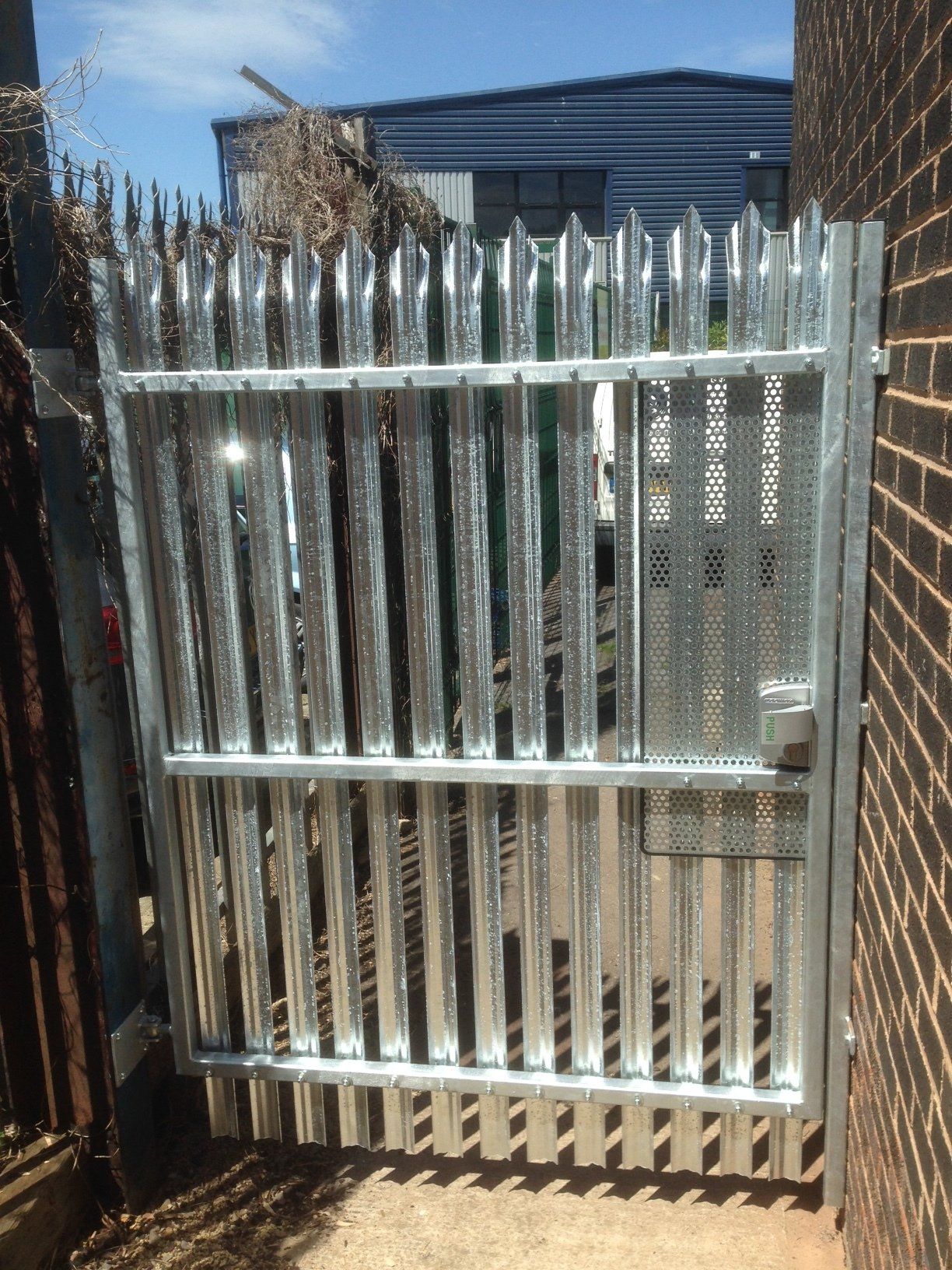Metal gate with vertical spikes and perforated panel, set in a brick wall.