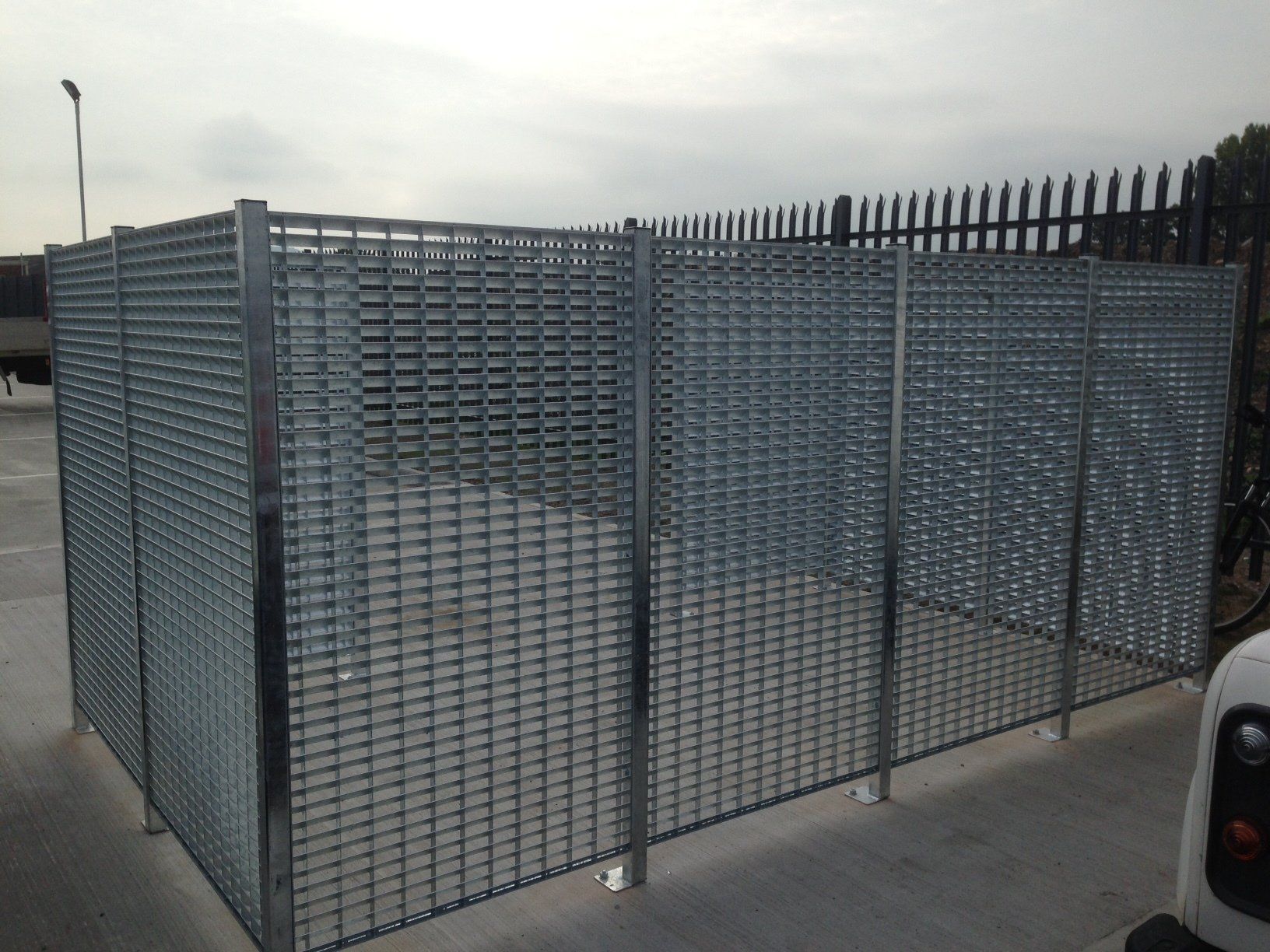Metal grid enclosure outdoors, likely for storage. Grey metal posts and mesh. Concrete ground and overcast sky.