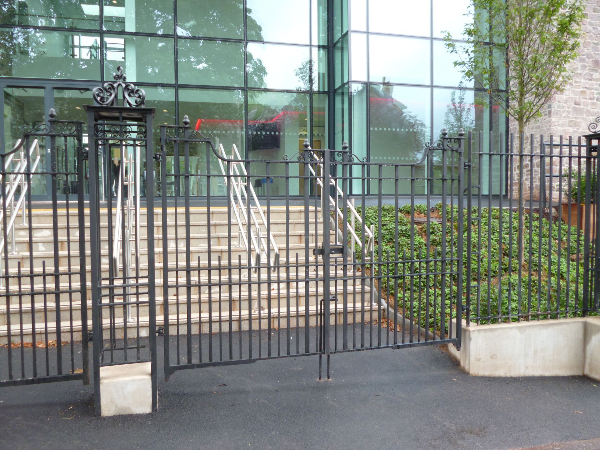Black wrought-iron gate in front of a modern building with glass windows, leading to stairs and a green hedge.