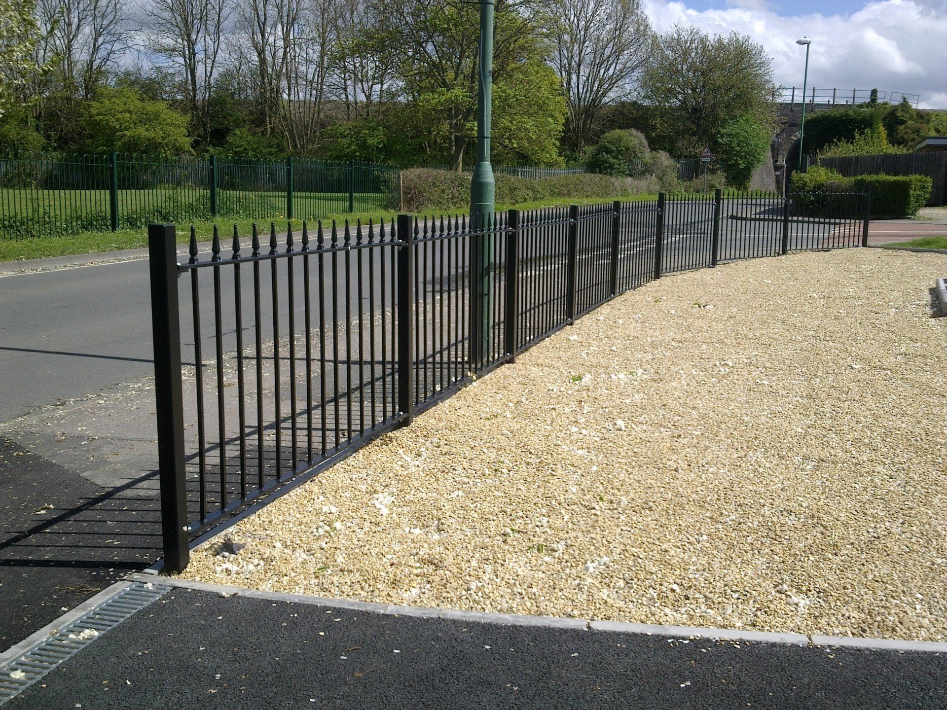 Black metal fence with pointed tops alongside gravel path and asphalt road.