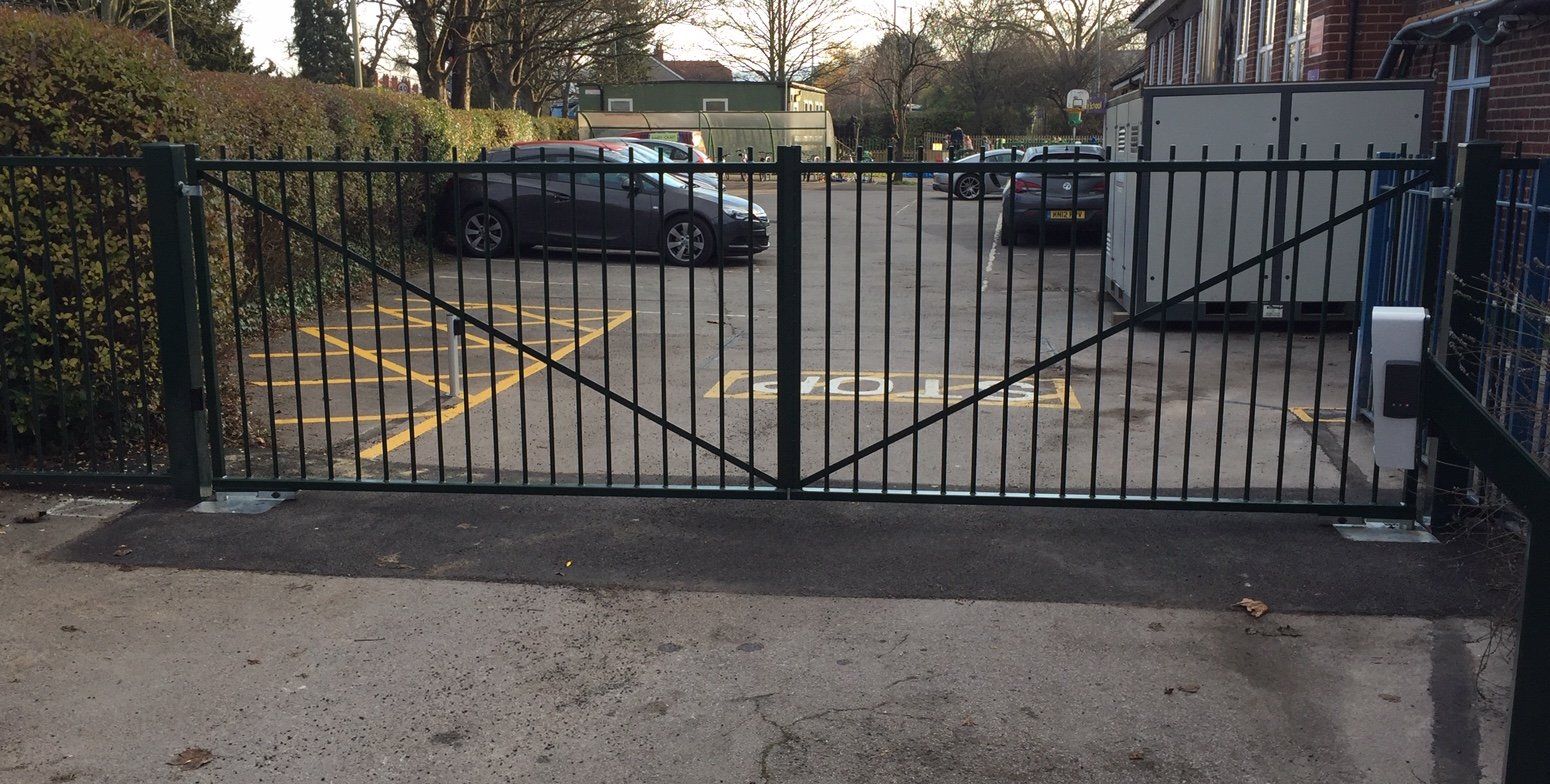 Black metal double gate across a parking lot with cars, next to a building and greenery.