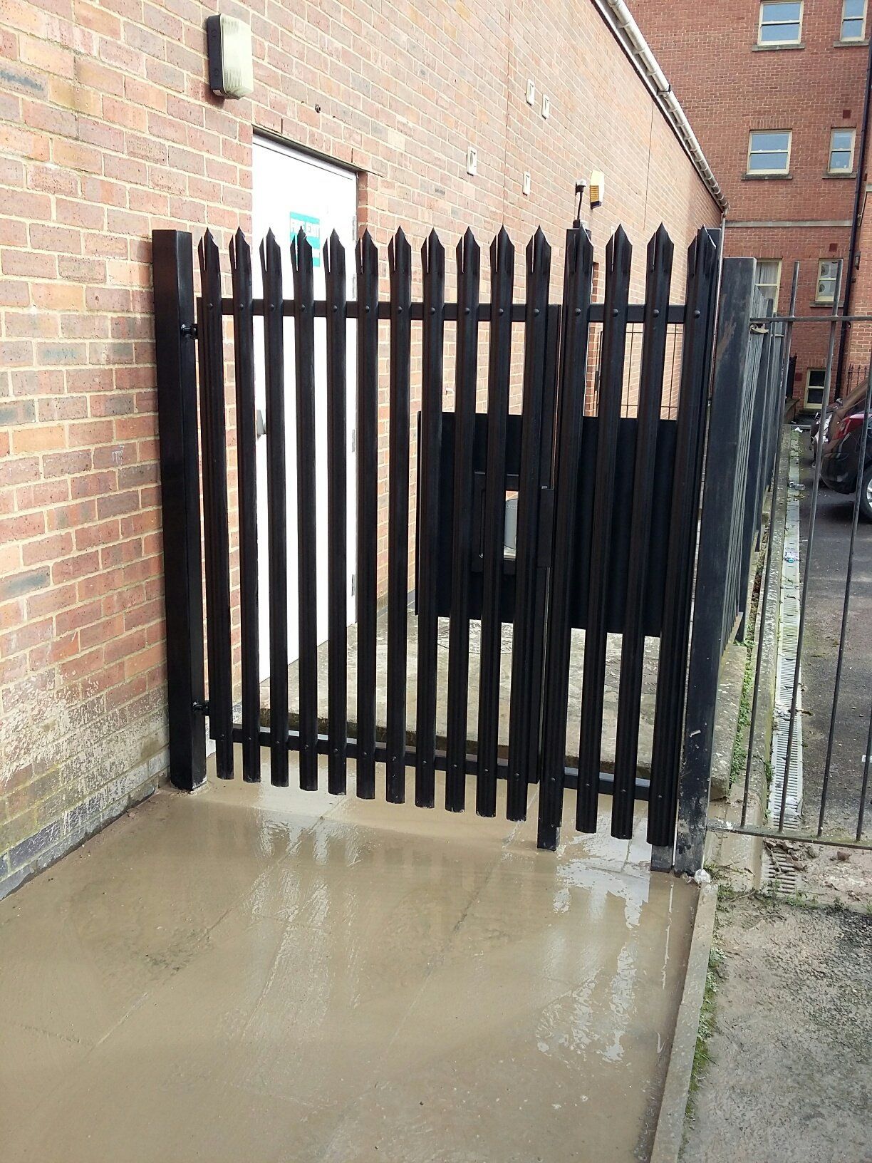 Black metal gate in a narrow space between a brick wall and a fence on a wet concrete surface.