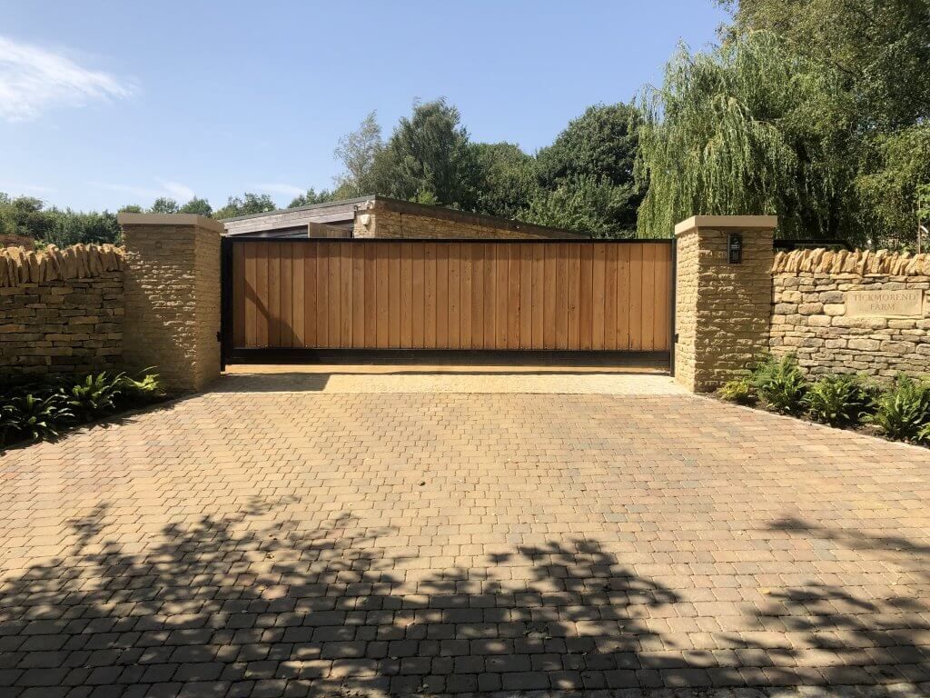 Wooden driveway gate set between stone pillars, leading to a home; brick paving.