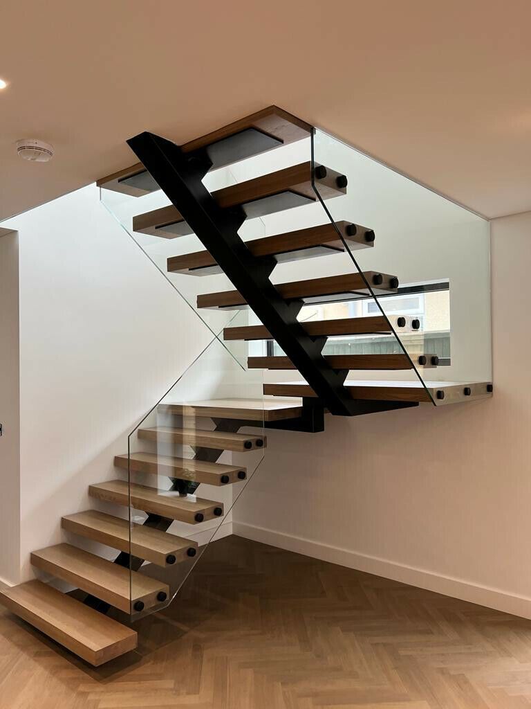 Modern wooden staircase with glass railing. Black metal spine, light wood steps, white walls, and wood floor.