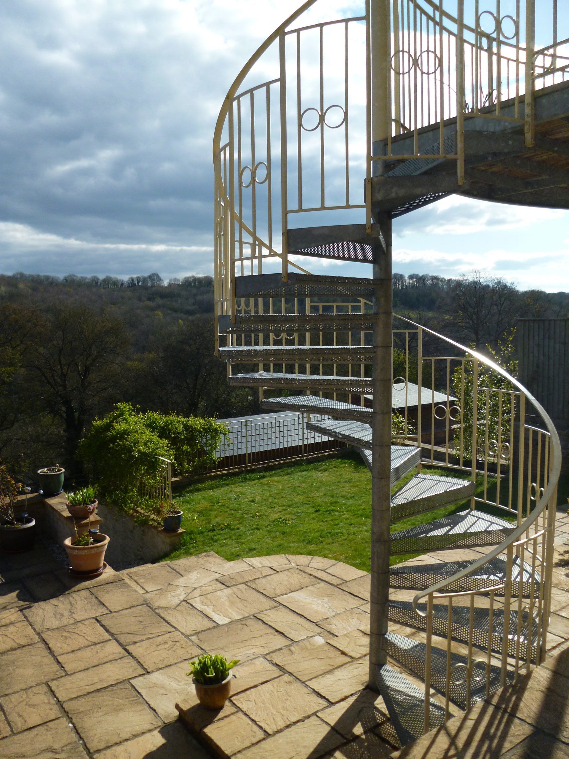 Spiral staircase outdoors with a metal railing and steps leading up to a platform, overlooking a grassy yard.