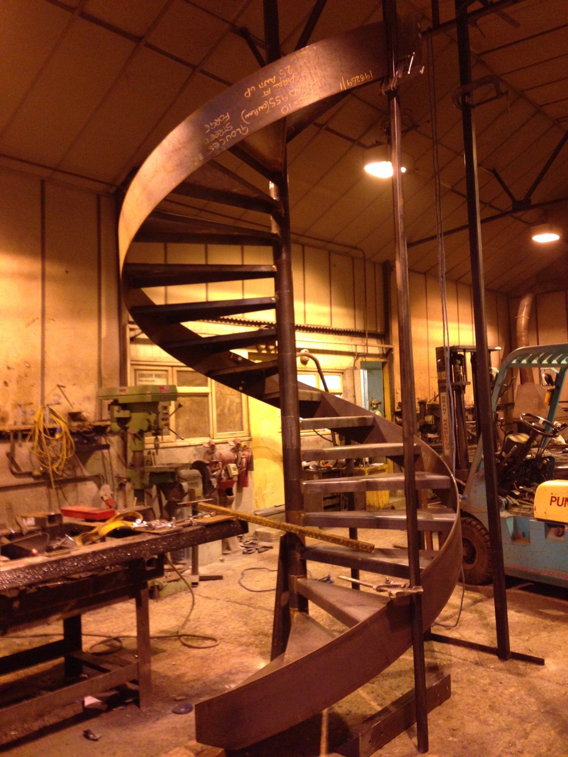 Spiral staircase made of metal in a workshop, dark brown against a neutral backdrop.