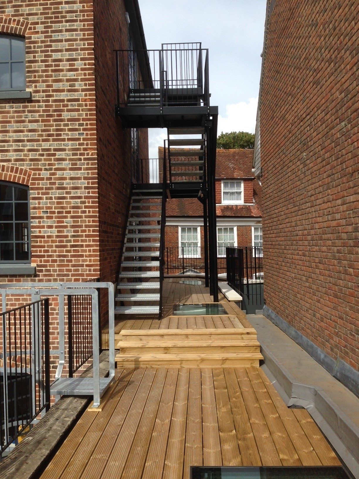A wooden deck with a black metal fire escape between two brick buildings.