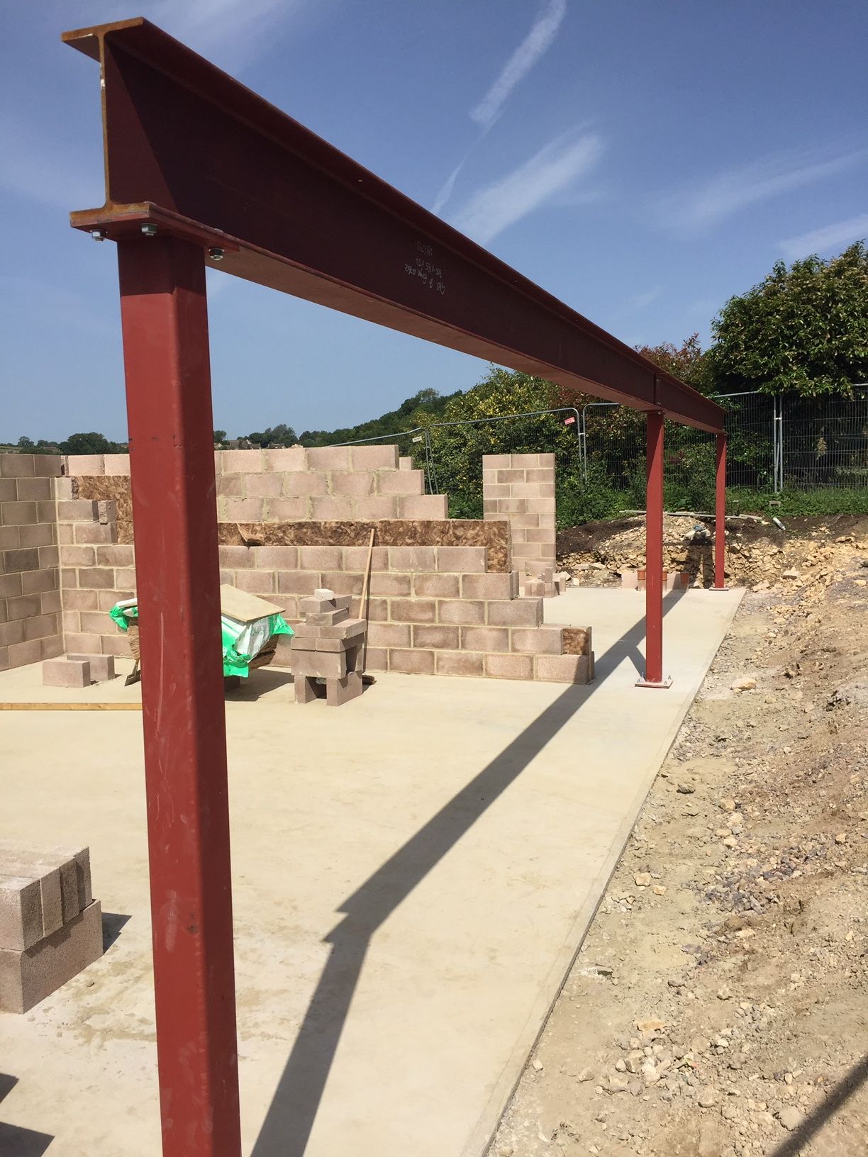 Red steel beams forming a frame over a concrete slab at a construction site with brickwork and a sunny sky.
