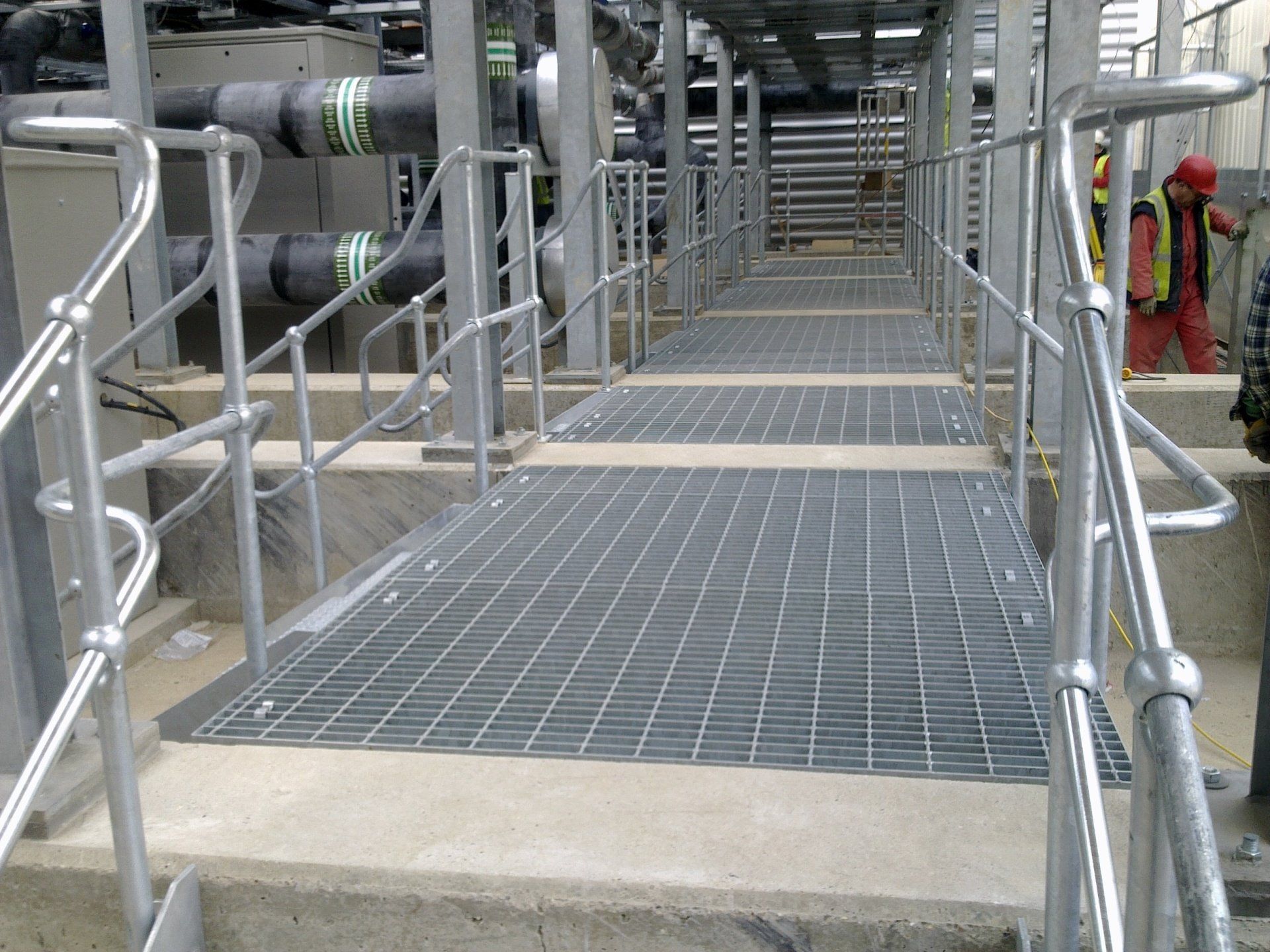 Metal grate walkway with railings, in an industrial setting. A worker in the background.