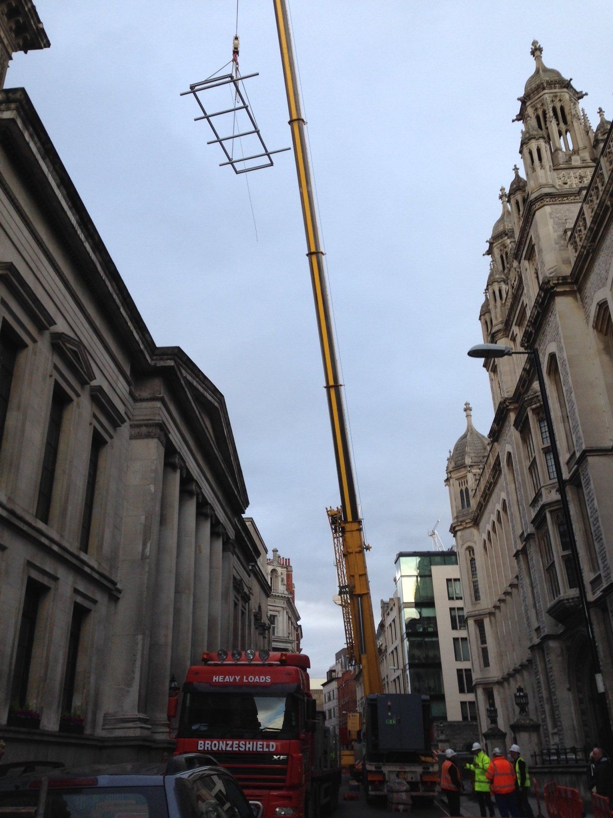 Crane lifting a rectangular structure in a city street. Buildings line both sides. Cloudy sky overhead.