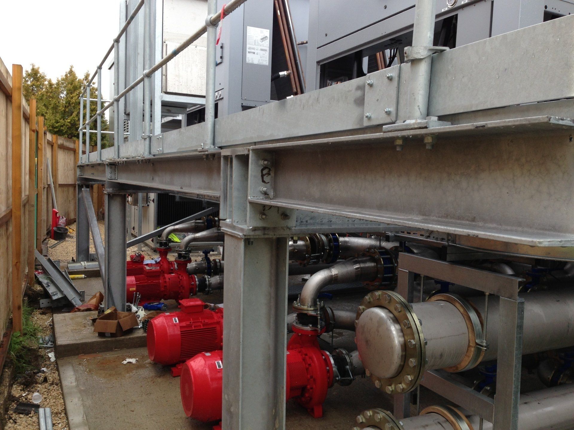 Metal platform with railing next to machinery, including red pumps, pipes, and a fence.